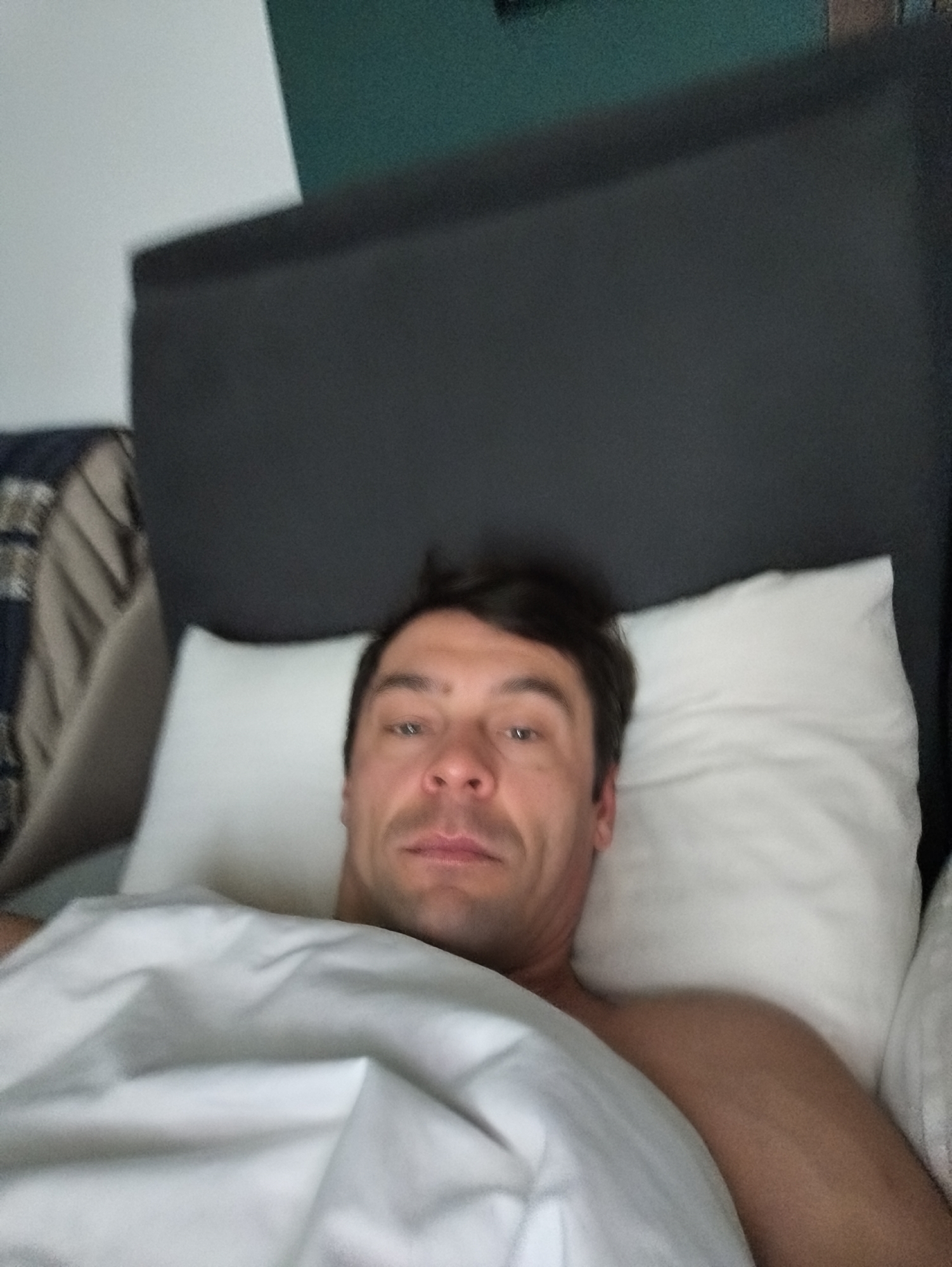 A man is lying in a bed. He is under a white sheet and has his head resting on a white pillow. Behind the pillow is a gray headboard. Behind the headboard is a dark green wall. A throw blanket is visible on a sofa to the left. The location is not discernible from the image.