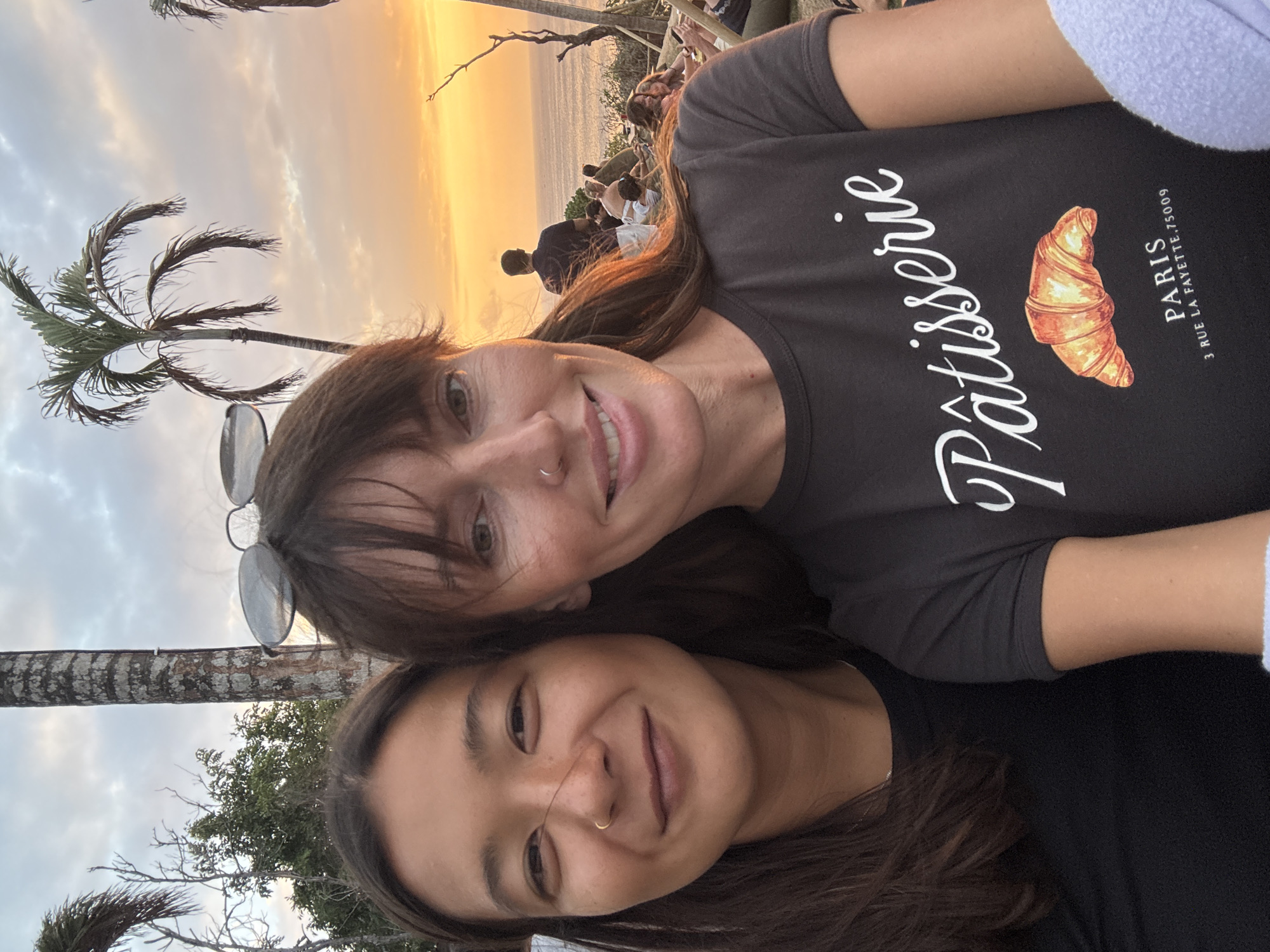 Two women are taking a selfie in front of a sunset. The woman on the left has long dark hair and is wearing a black t-shirt. The woman on the right has shoulder-length brown hair and is wearing a dark grey t-shirt with a graphic of a croissant and the words "Pâtisserie PARIS 3 RUE LAFAYETTE, 75009" on it. She has sunglasses perched on her head. The background features palm trees and a cloudy sky with an orange sunset. In the distance, there are other people sitting and looking towards the ocean. This appears to be an outdoor seating area with a view of the water. The location is likely a restaurant or bar in Kuta Selatan, Indonesia, known for its sunset views.