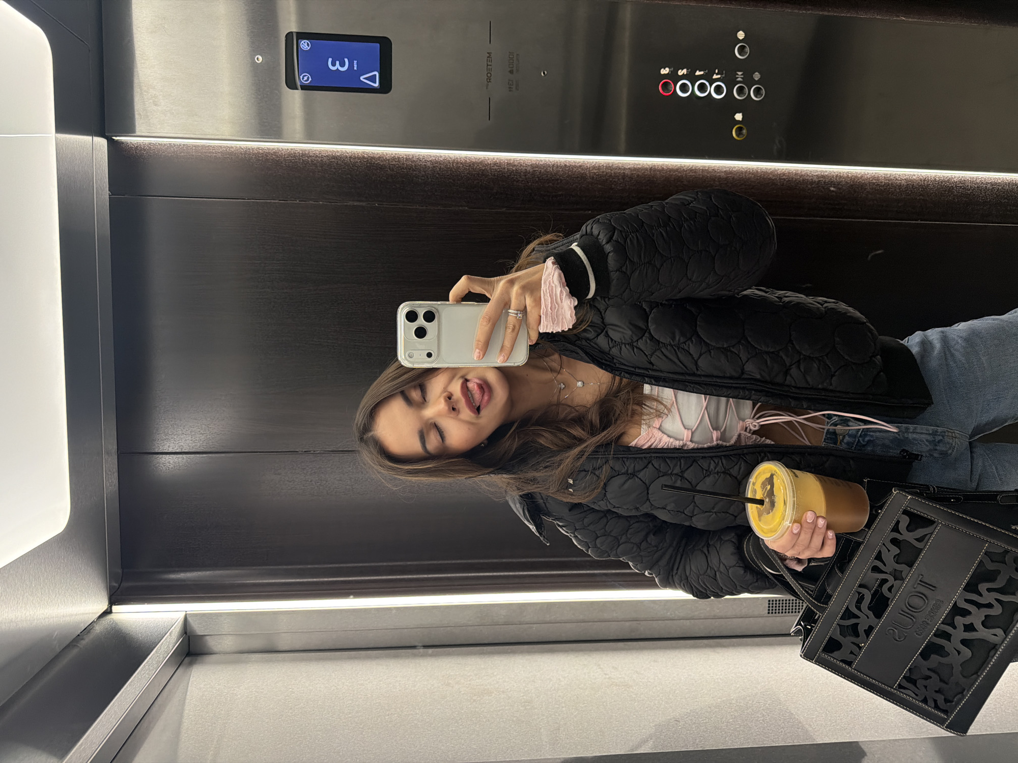 The image is a selfie taken by a young woman inside an elevator. She is wearing a black puffer jacket, a light-colored top with pink strings, and blue jeans. She's holding a drink in a plastic cup with a straw in one hand and a smartphone in the other, using it to take the picture. A black tote bag with cut-out patterns is also visible. The woman is sticking her tongue out. The elevator is modern with a digital display showing the floor number "3". There is a panel with elevator buttons. The walls of the elevator are a combination of light gray and dark wood-like paneling with a strip of light at the bottom. It is unknown if the location is in the city of Krasnoyarsk, Russia.
