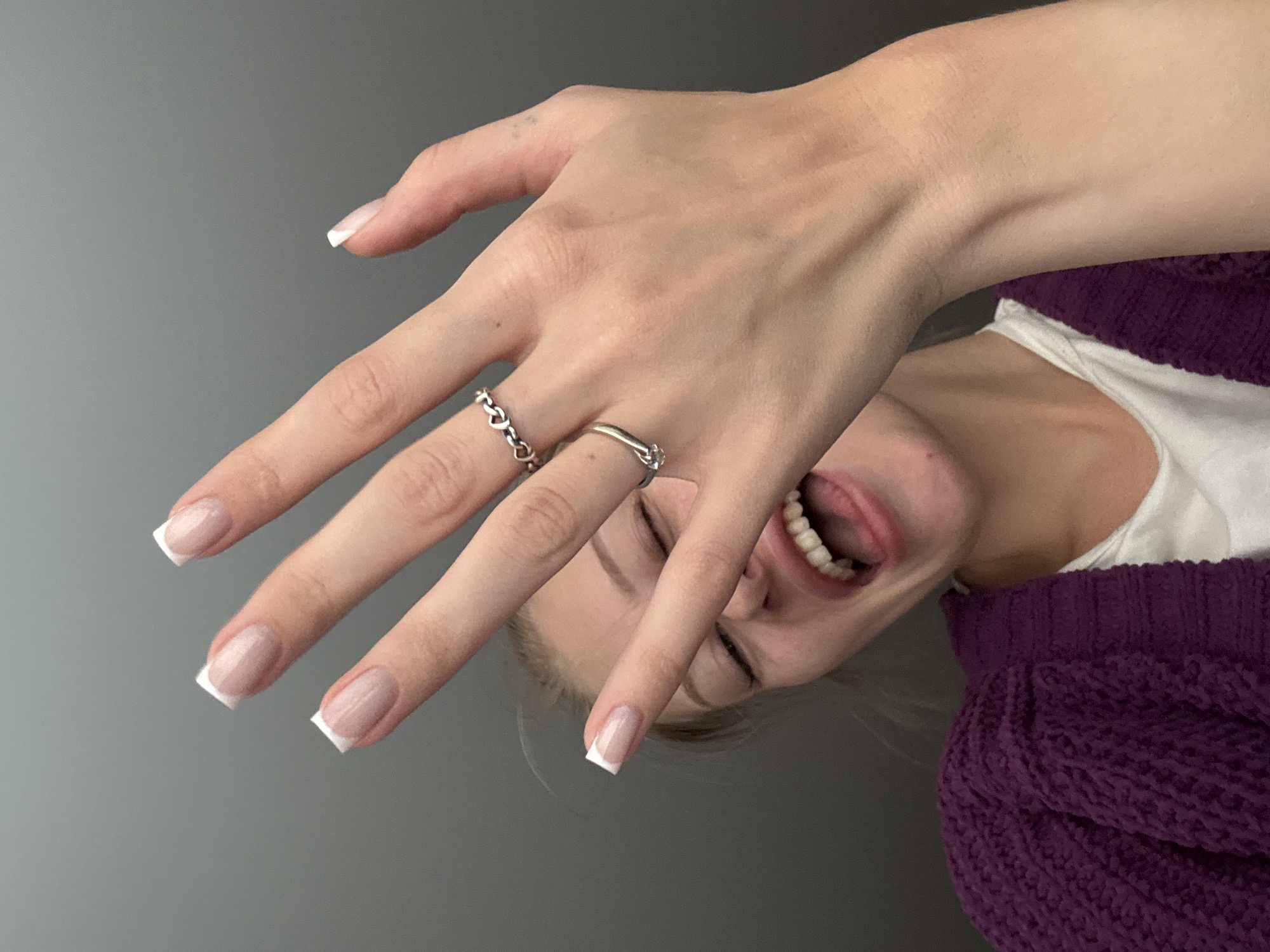 The image shows a young woman with her hand raised in front of her face, obscuring her eyes. She is wearing a purple knitted cardigan over a white top. Her fingernails are painted with a French manicure. She is wearing two rings on her fingers: one thin, silver ring with a heart design and a solitaire diamond ring. Her mouth is open, and her tongue is visible, suggesting a playful or expressive pose. The background is a plain, gray wall, consistent with an indoor setting. There is no indication of the city of Perm, Russia, or any other specific location.