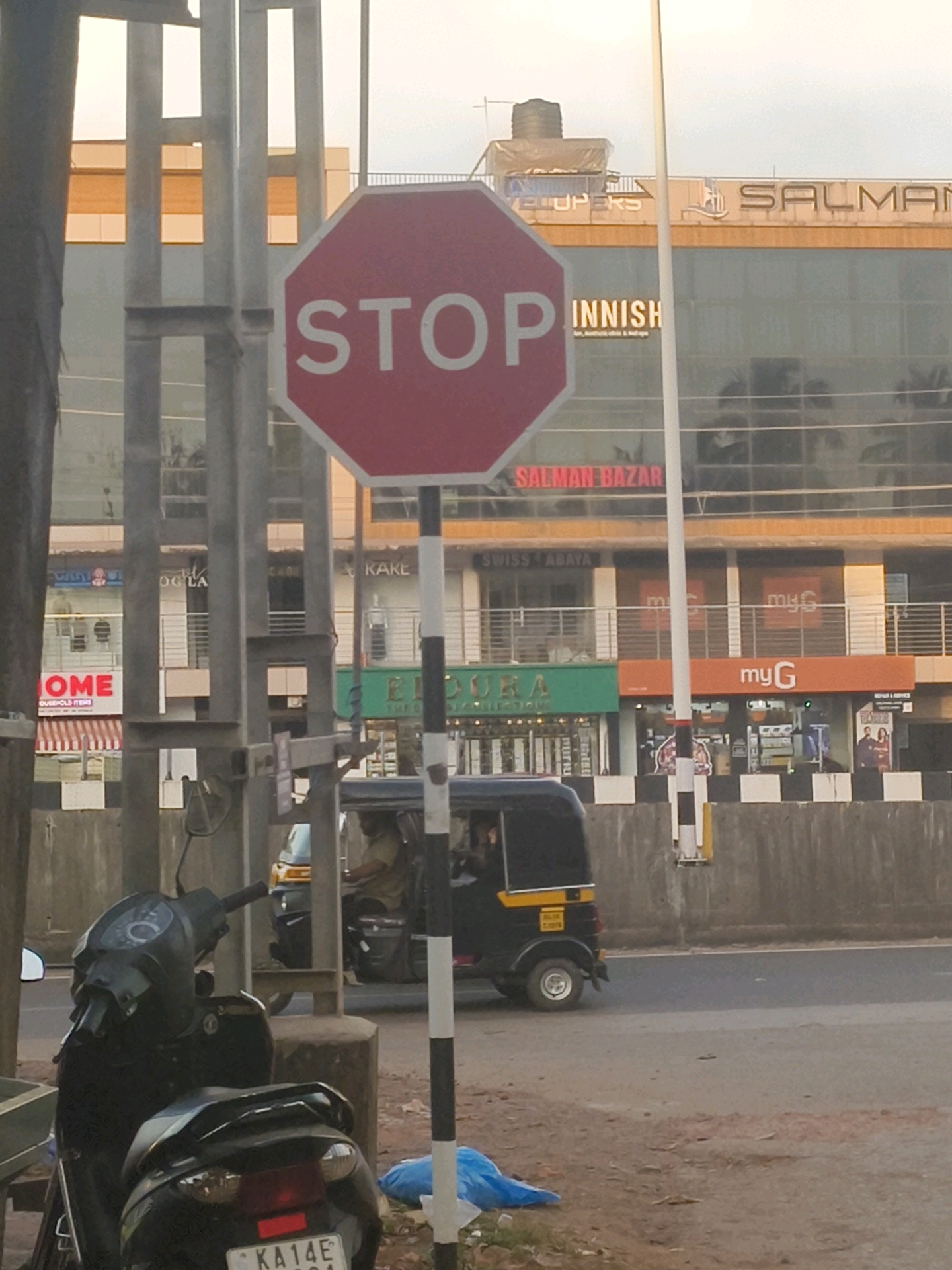 The image depicts a street scene in Mangalpady, India, dominated by a red octagonal stop sign in the foreground. Behind the sign, a multi-story building features large glass windows and several storefronts with visible signage. Store names include "SALMAN BAZAR," "INNH," "SWISS ABAYA," "my G," and "HOME HOUSEHOLD ITEMS." A black auto-rickshaw with a yellow stripe is visible on the road, and two motorcycles are parked on the left side of the frame. A concrete barrier runs along the side of the road. The sky is overcast.