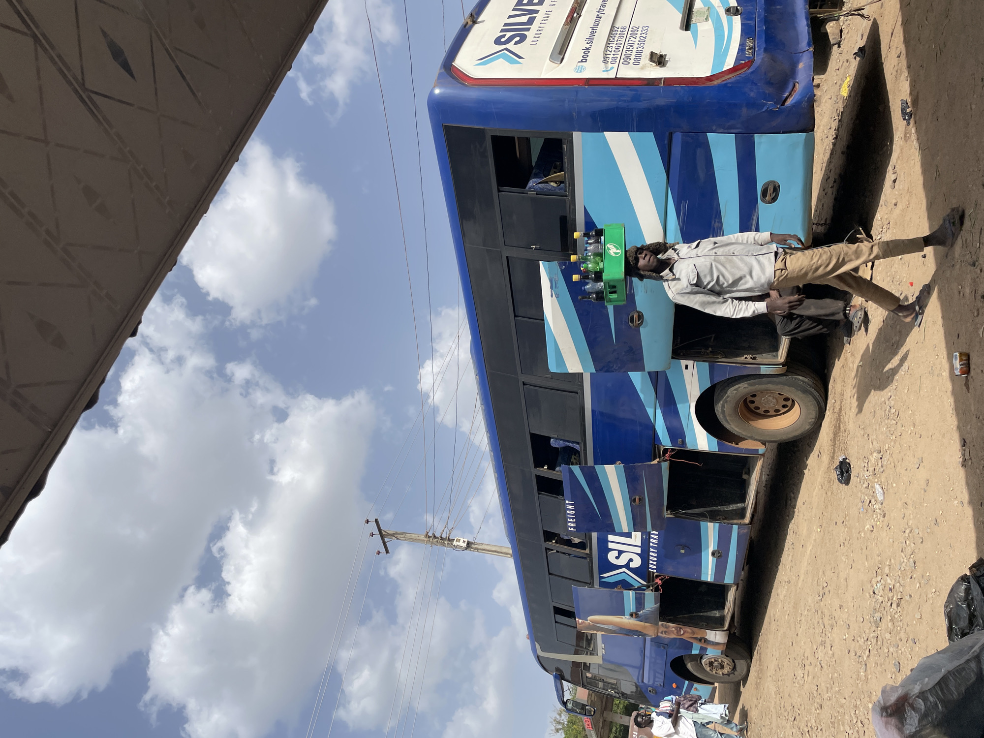 This daytime image, set in Jos, Nigeria, captures a vibrant blue and white long-distance bus parked on a dusty, unpaved roadside under a bright, partly cloudy sky. The bus is decorated with prominent branding including "SILVER LUXURY TRAVEL" and "SIL FREIGHT" along its side. Several cargo compartments on the lower side of the bus are open, revealing dark interiors.
A man, wearing a light-colored collared shirt, tan trousers, sandals, and a dark, possibly fur-trimmed hat, stands next to the bus. He is holding a green plastic crate filled with several plastic bottles of beverages, suggesting he is a vendor. Another person is partially visible near the front wheel of the bus. In the background, a wooden utility pole supports multiple power lines stretching across the clear blue sky. The ground is uneven, littered with small rocks and debris, with shadows indicating bright sunlight.
Visible text on the bus includes "SILVER LUXURY TRAVEL", its website "book.silverluxurytravels.com", and several Nigerian phone numbers: "09123164602", "08106078068", "09034072092", and "08083502333". The words "FREIGHT" and "SIL LUXURY TRAV" are also legible on the side.