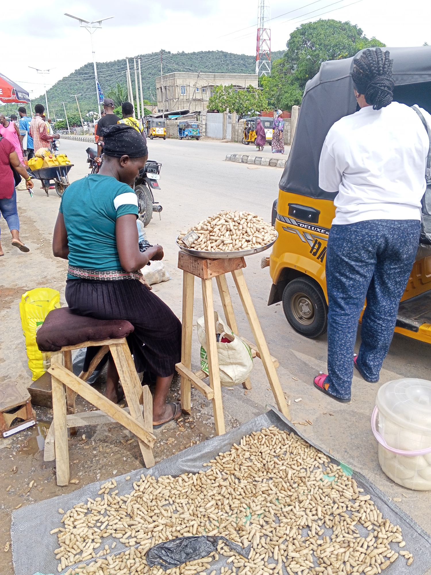 This image captures a street scene in Zango-ndaji, Nigeria, featuring a market stall with peanuts displayed for sale. In the foreground, a large mat covers the ground, laden with a substantial quantity of peanuts. To the right of the mat is a white plastic bucket with a pink rim, possibly used for storage or display.
A woman wearing a teal t-shirt and a dark skirt is seated on a wooden stool, positioned next to a stand holding a metal tray filled with peanuts. She appears to be engaged in an activity related to the peanuts. A yellow plastic bag is visible behind her. A wooden stand supports the tray of peanuts, and a white cloth bag hangs from it.
In the midground, a yellow motorized tricycle, commonly known as an "okada" or "Keke Napep," is parked. A woman in a white t-shirt and patterned blue pants stands next to it, facing away from the camera. The tricycle has a black canopy.
Further in the background, various individuals are seen near motorcycles and other vehicles on a paved road. The surroundings include buildings under construction, a tall red and white communication tower, and a lush green hillside in the distance. Power lines are visible overhead. The overall environment suggests an active street market or commercial area.