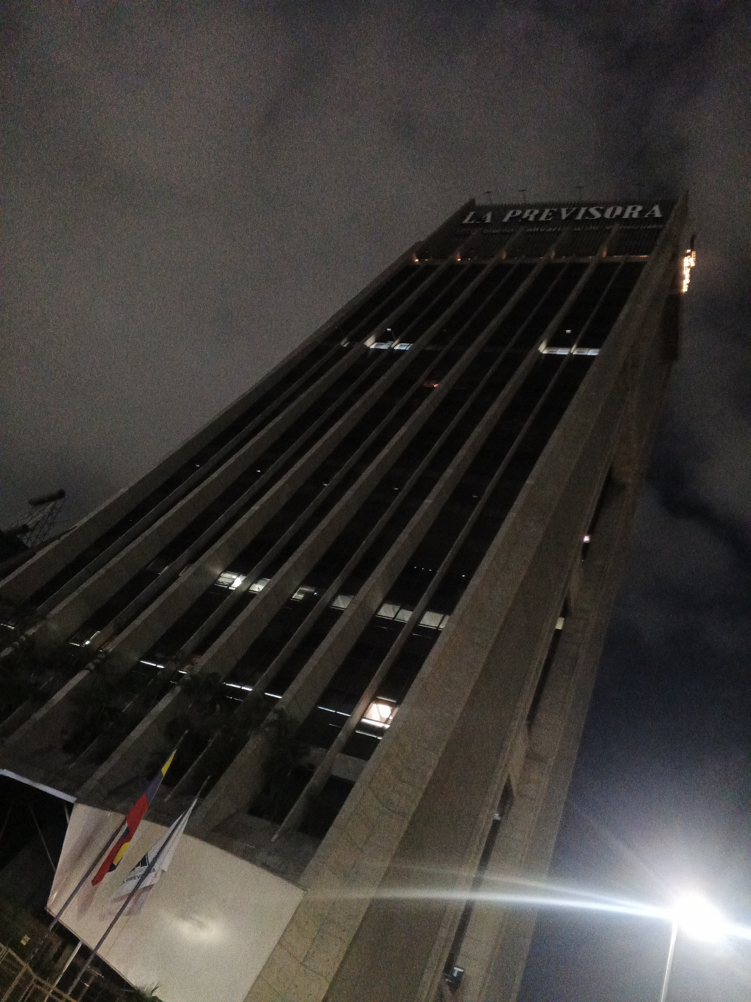 An upward angled, nighttime photograph shows the exterior of a tall, modern skyscraper in Caracas, Venezuela. The building features a repeating pattern of angled concrete fins and dark windows. The name "LA PREVISORA" is visible in large white lettering on the top of the building. The sky is cloudy and dark. A bright light, possibly a streetlamp, flares across the bottom right of the image. At the base of the building, two flags are visible, one of which appears to be the flag of Colombia. The scene is illuminated by artificial lights from within the building and the exterior light source.