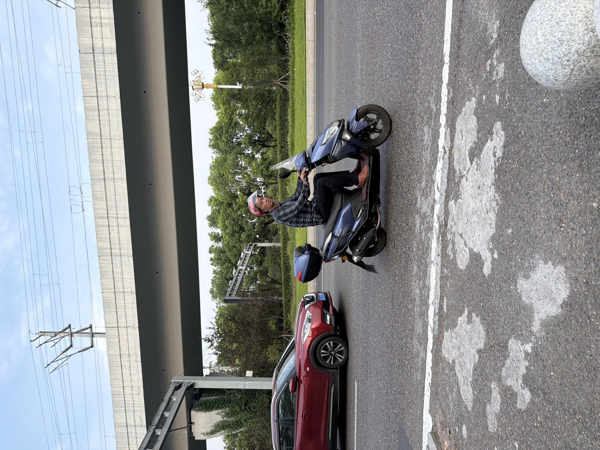 A man wearing a red helmet and a plaid shirt rides a blue scooter on an asphalt road in Jiangjin, China. The man is looking towards the viewer. A red SUV is visible to the left of the scooter. In the background, trees and a street lamp can be seen. Above, there is a large overpass with power lines. The sky is partly cloudy.