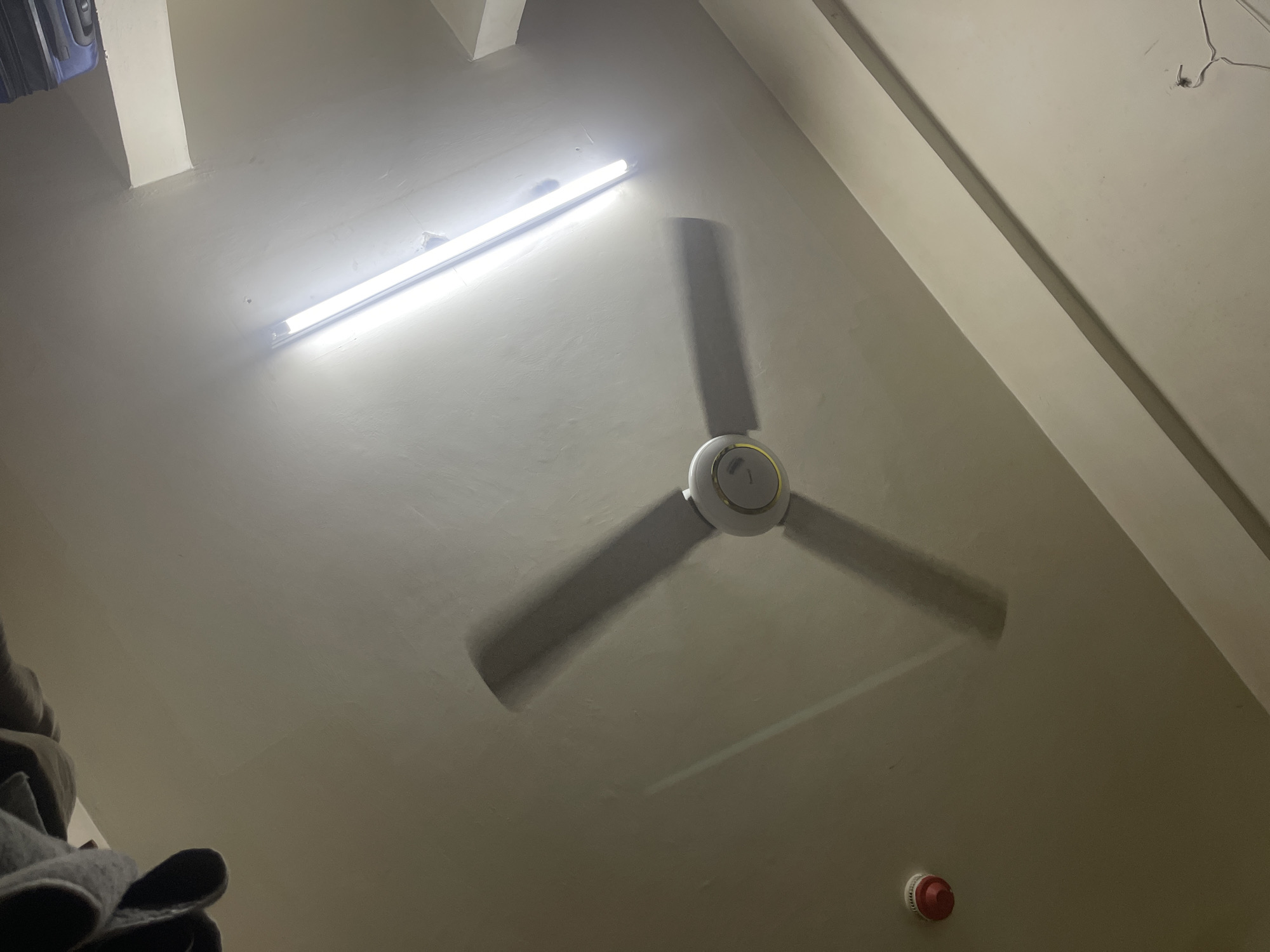 The image shows an upward view of a room's ceiling. A fluorescent light fixture illuminates the left side of the ceiling. A three-blade ceiling fan is mounted centrally. A smoke detector is visible near the bottom right corner. Part of what appears to be a suitcase or bag is visible in the top left corner, and gray clothing is visible in the bottom left corner. The walls appear to be painted a light color. There is no indication of location.
