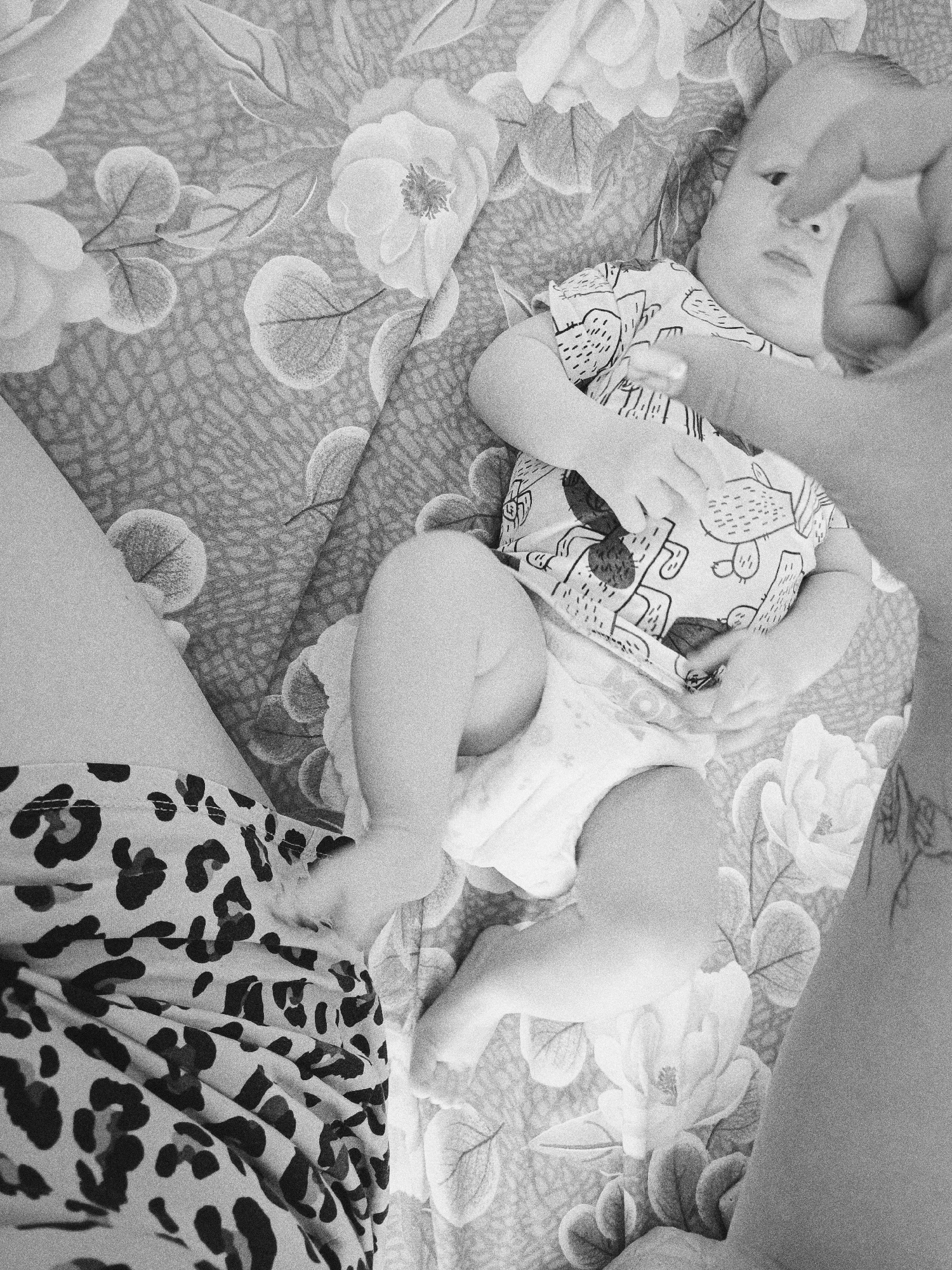 A black-and-white image shows a baby lying on a floral-patterned blanket, with an adult partially visible on either side. The baby is wearing a patterned onesie and a diaper. The adult on the left is wearing animal-print pants, while the adult on the right has a tattoo visible on their arm. The location is not discernible from the image.