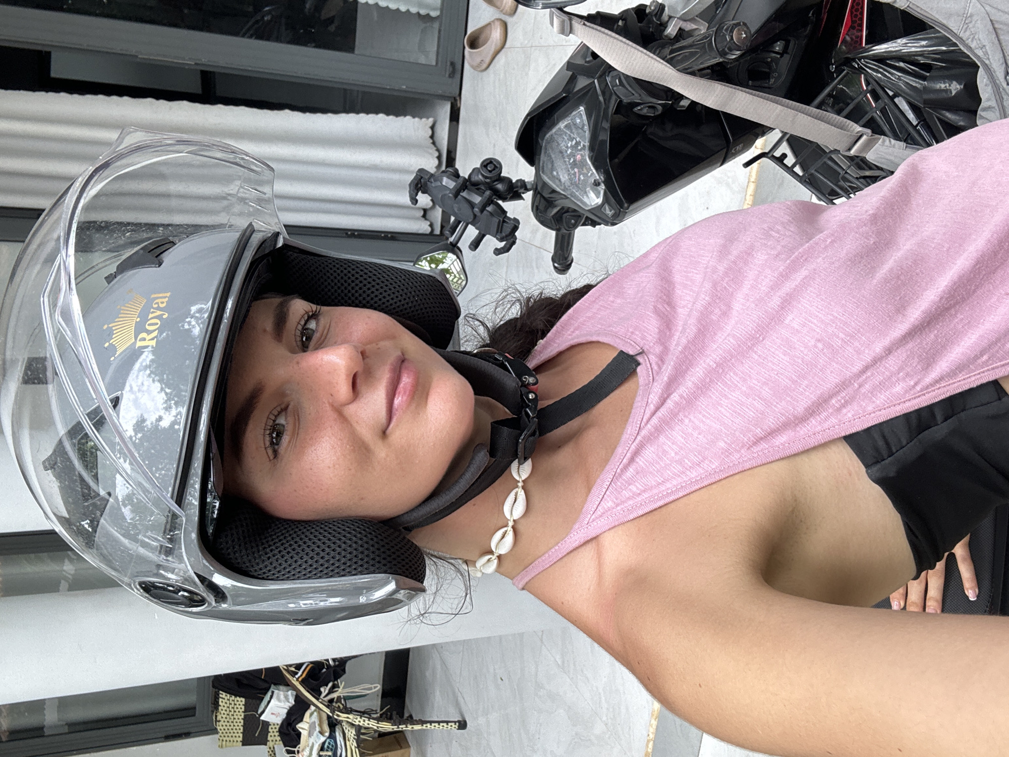 A close-up, rotated portrait shows a woman wearing a grey motorcycle helmet with a clear visor, looking directly at the camera with a soft smile. Her dark hair is visible, and she wears a light pink sleeveless top over a black undergarment. A necklace made of white cowrie shells adorns her neck.
The helmet features a gold "Royal" brand name with a crown logo on its side. To the right and behind her, parts of a black motorcycle or scooter are visible, including its handlebars, a headlight, and a phone holder mounted on the dashboard. A grey strap, possibly from luggage, is draped over the motorcycle. A black basket or rack is also seen towards the front of the vehicle.
The background suggests an outdoor setting, likely a paved area next to a building. A pair of light brown slip-on shoes rest on the ground near the motorcycle. Behind the woman's head, a glass door or window with white horizontal blinds or shutters is visible. Some clothes or fabric are piled on the ground in the lower-left corner. The bright, natural lighting indicates it is daytime. The overall scene suggests the woman is in Mũi Né, Vietnam, either preparing for or having just completed a scooter ride.