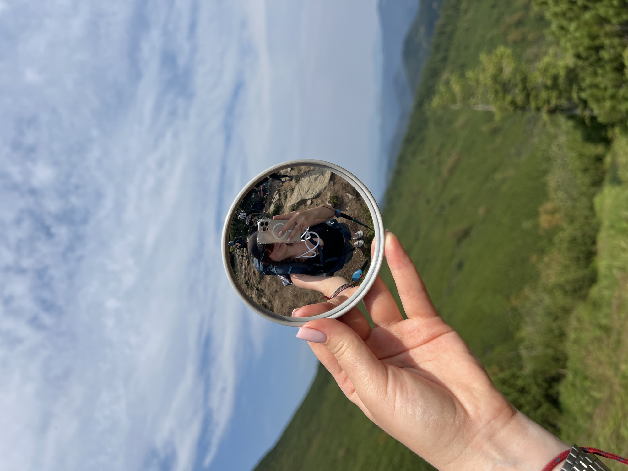 In the foreground, a person's left hand, with light pink painted fingernails, holds a round mirror. The mirror reflects a selfie of a woman wearing sunglasses, a black top, and a backpack. She is holding a smartphone in her right hand, also visible in the reflection. Behind the hand and mirror, a lush green, undulating mountain landscape stretches into the distance under a cloudy blue sky. The scene appears to be a scenic location, possibly in the Carpathian Mountains near Vorokhta, Ukraine, given the natural, mountainous terrain and the presence of hikers.