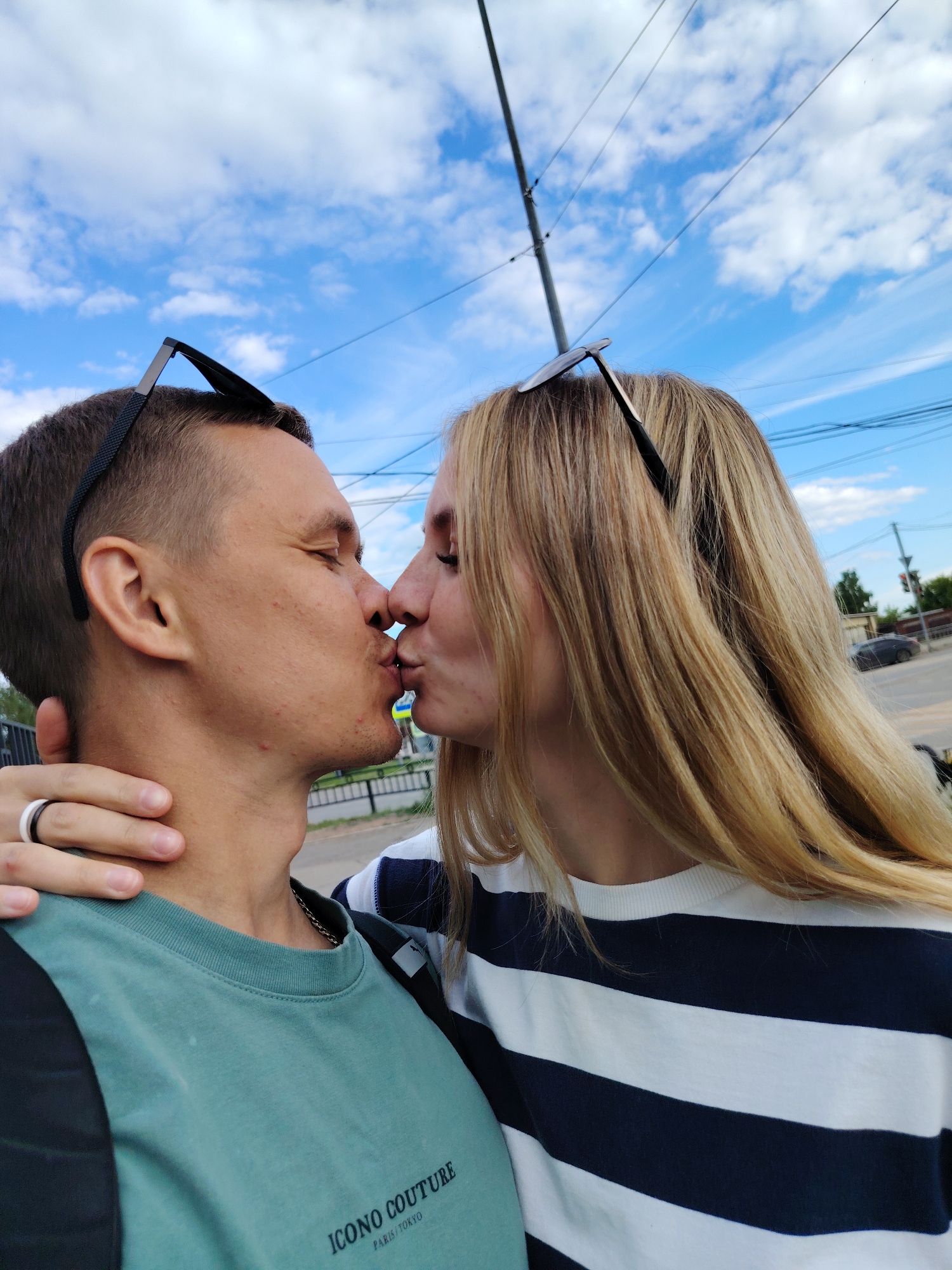 A vertical outdoor photograph captures two individuals kissing on a clear, sunny day in Chaikovsky, Russia. The person on the left, with short dark hair and sunglasses pushed onto their forehead, wears a teal-colored long-sleeve shirt with a black shoulder panel, featuring the text "ICONO COUTURE PARIS/TOKYO". A silver chain is visible around their neck, and a black and white ring is on their left ring finger. Their eyes are closed. The person on the right, with long blonde hair and sunglasses also pushed onto their head, wears a white shirt with horizontal navy blue stripes. Their eyes are also closed. Their left hand is gently placed on the neck/upper shoulder of the other individual. Their lips are pressed together in a kiss. The sky is bright blue with scattered white cumulus clouds. In the background, a utility pole extends upwards, supporting multiple power lines that traverse the upper portion of the frame. A black metal fence is partially visible behind the left individual. Further back, a road features a partially obscured pedestrian crossing sign, and a dark sedan is visible. Distant greenery and indistinct buildings line the horizon.