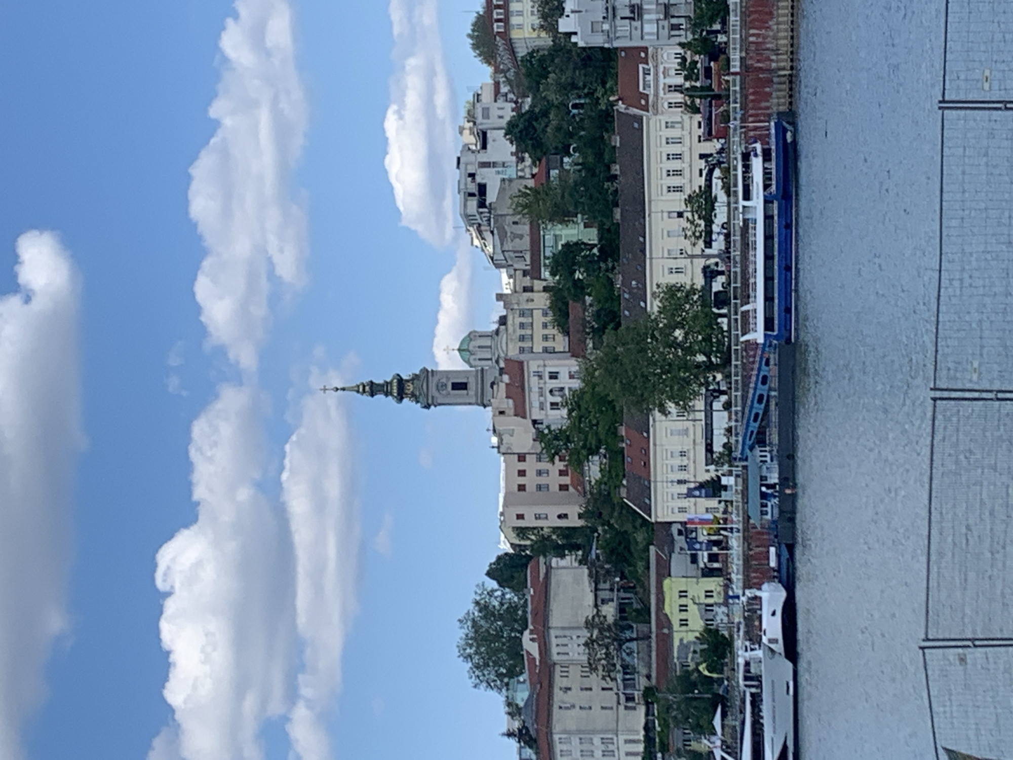 The image displays a picturesque daytime scene of a city situated along a river, identified as Belgrade, Serbia. Under a clear blue sky dotted with scattered white cumulus clouds, a vibrant urban landscape rises from the water's edge.
The city, positioned on the left side of the frame, features a dense collection of multi-story buildings, predominantly light-colored with red-tiled roofs, characteristic of European architecture. A prominent landmark is a grand church or cathedral, distinguished by its tall, ornate bell tower crowned with a spire and cross, and a striking green dome. Lush green trees are interspersed among the buildings, providing natural elements within the urban setting.
To the right, a wide body of calm, grey-blue water, likely a river, stretches across the frame. Several boats are docked along the riverbank. A notable vessel is a long, blue and white boat, possibly a tour boat or ferry, moored prominently. Another smaller white boat is also visible further along the embankment. The riverfront is lined with a concrete or stone embankment, and some metallic structures, possibly part of a dock or railing, are visible on the far right.
The scene is bathed in clear daylight, indicating either morning or afternoon. No specific human activity is evident, and there is no discernible text on any of the visible objects or buildings. The overall atmosphere is tranquil, showcasing a scenic view of a historic riverside city under fair weather conditions.
