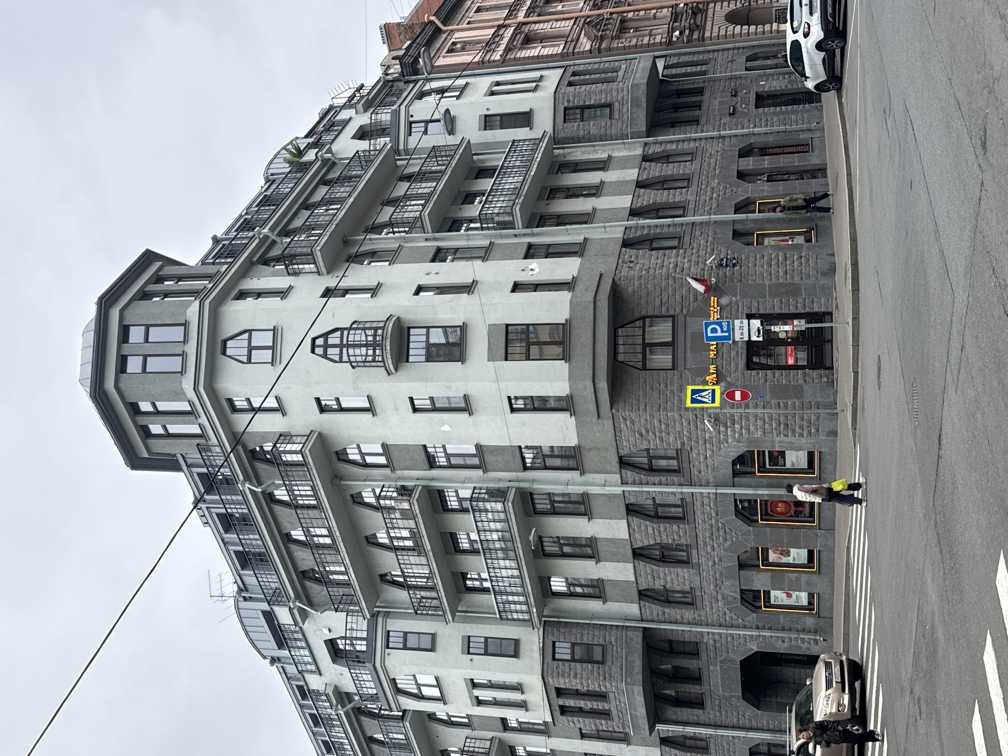 The image depicts an urban scene in Saint Petersburg, Russia, featuring a distinctive building with a modern architectural style. The building is characterized by a pale gray facade, large windows, and cantilevered balconies with black railings. A portion of the building's lower level is clad in dark gray stone. On the right side of the building, at street level, are signage, including parking signs, and entrances to what appear to be commercial establishments. Pedestrians are visible on the crosswalk, including a person wearing a light-colored jacket and carrying a yellow bag. Cars are parked along the street, and telephone wires extend across the upper portion of the frame. The sky is overcast, suggesting a cloudy day.