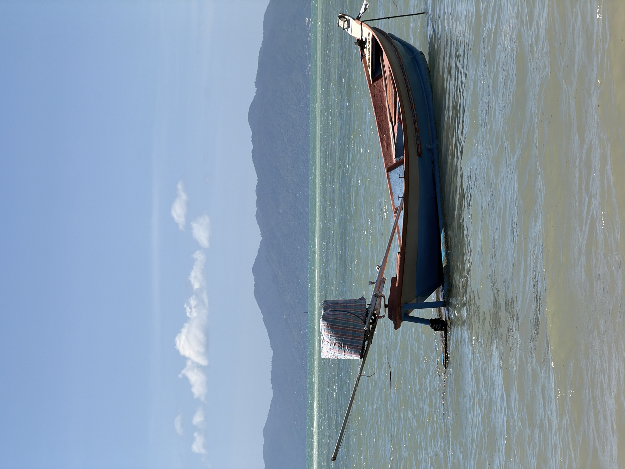 The image depicts a small, blue and red fishing boat floating on calm, slightly murky water. The boat is tethered and anchored. A large, checkered bundle is secured to a metal pole extending from the boat. In the background, a hazy mountain range with some visible structures along its base stretches across the horizon. The sky is a clear, light blue with a few wispy clouds. The scene is set in the Ko Samui District of Thailand, likely near a coastal area. There are no people visible in the image. The environment appears to be a sunny day with gentle waves.