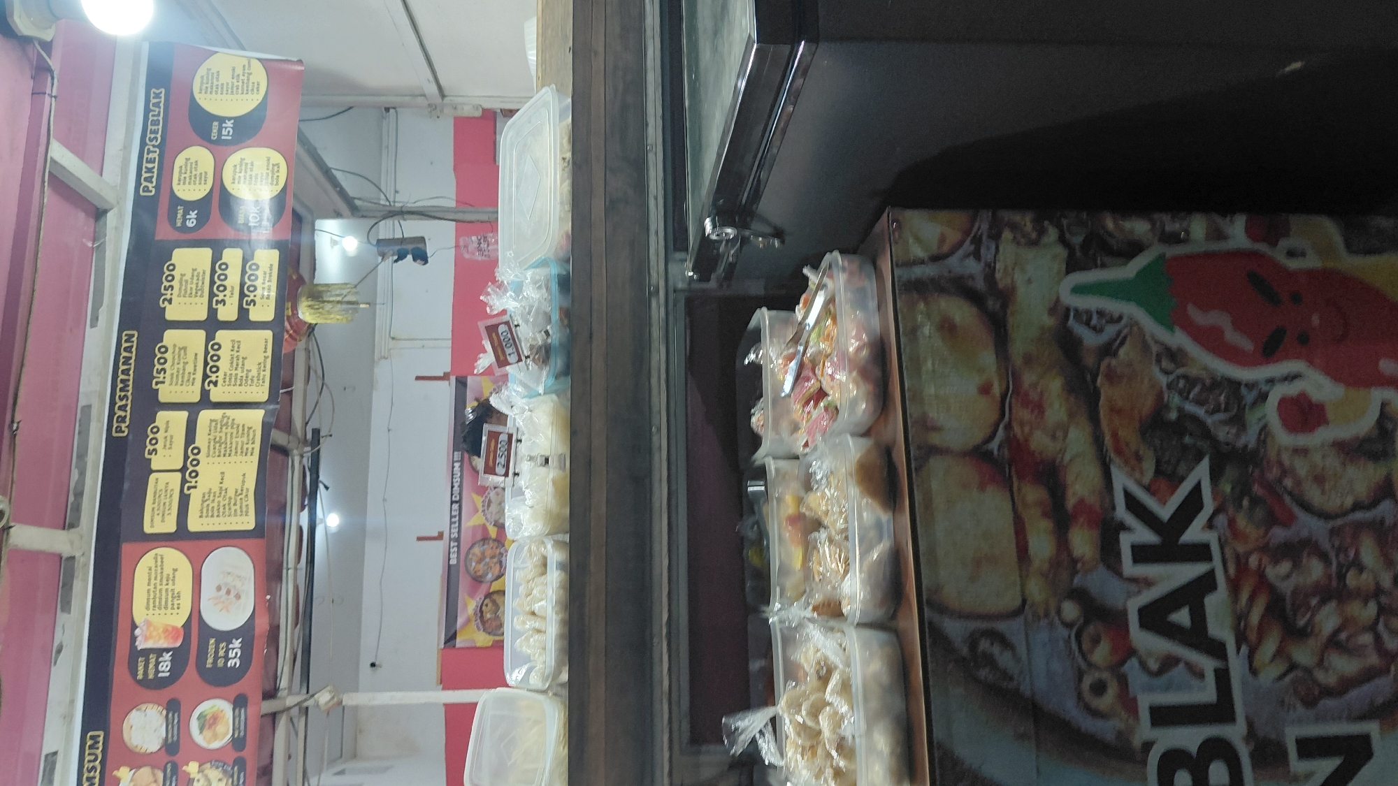 The image shows a street food stall in Bantarsoka, Indonesia. The stall features a menu board with various food items and prices, including "Prasmanan" (buffet-style food) and "Paket Seblak" (seblak meal package). Various plastic containers filled with food are displayed on shelves. The stall also has a prominent sign with the word "BLAK" and a cartoon chili pepper illustration. The background includes the ceiling of the stall with lighting fixtures and some electrical wiring.