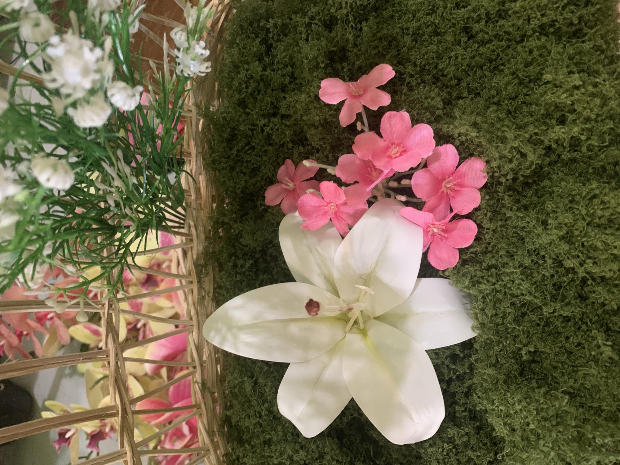 The image depicts a close-up, overhead view of artificial flowers arranged on a textured green surface, likely moss or a similar craft material. In the foreground, a large, white, multi-petaled flower, resembling a lily, is prominent. To its right, a cluster of smaller, bright pink flowers with five petals each are arranged. Behind these flowers, the texture of the green surface is visible, appearing dense and slightly ruffled. In the background, a woven, light brown basket structure is present, holding more artificial foliage and flowers in shades of pink and yellow. The lighting is even, suggesting an indoor setting. There are no people or discernible landmarks of Kuala Pembuang, Indonesia visible in this image.