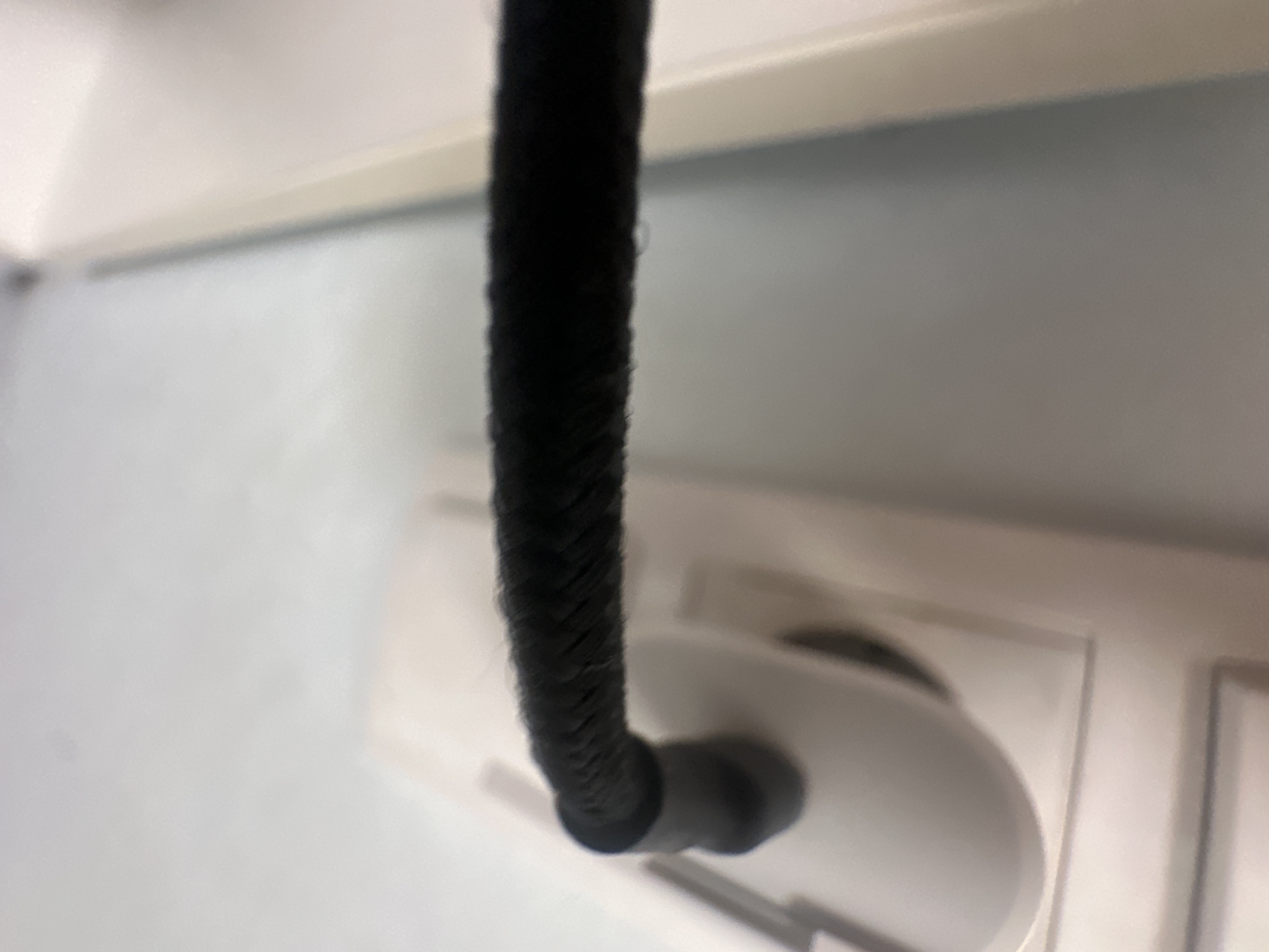 A black braided cable is plugged into a white electrical outlet on a light blue wall. The outlet is a standard European type with two circular holes. The cable appears to be made of a woven fabric material and is bent at an angle as it exits the outlet. The image is a close-up shot, with the focus primarily on the cable and the outlet. The background is a light blue wall, and the foreground is a white surface, possibly a shelf or table. No people or other identifying features are visible in the image. The scene is mundane and does not appear to be related to any specific event or location in Shumen, Bulgaria.