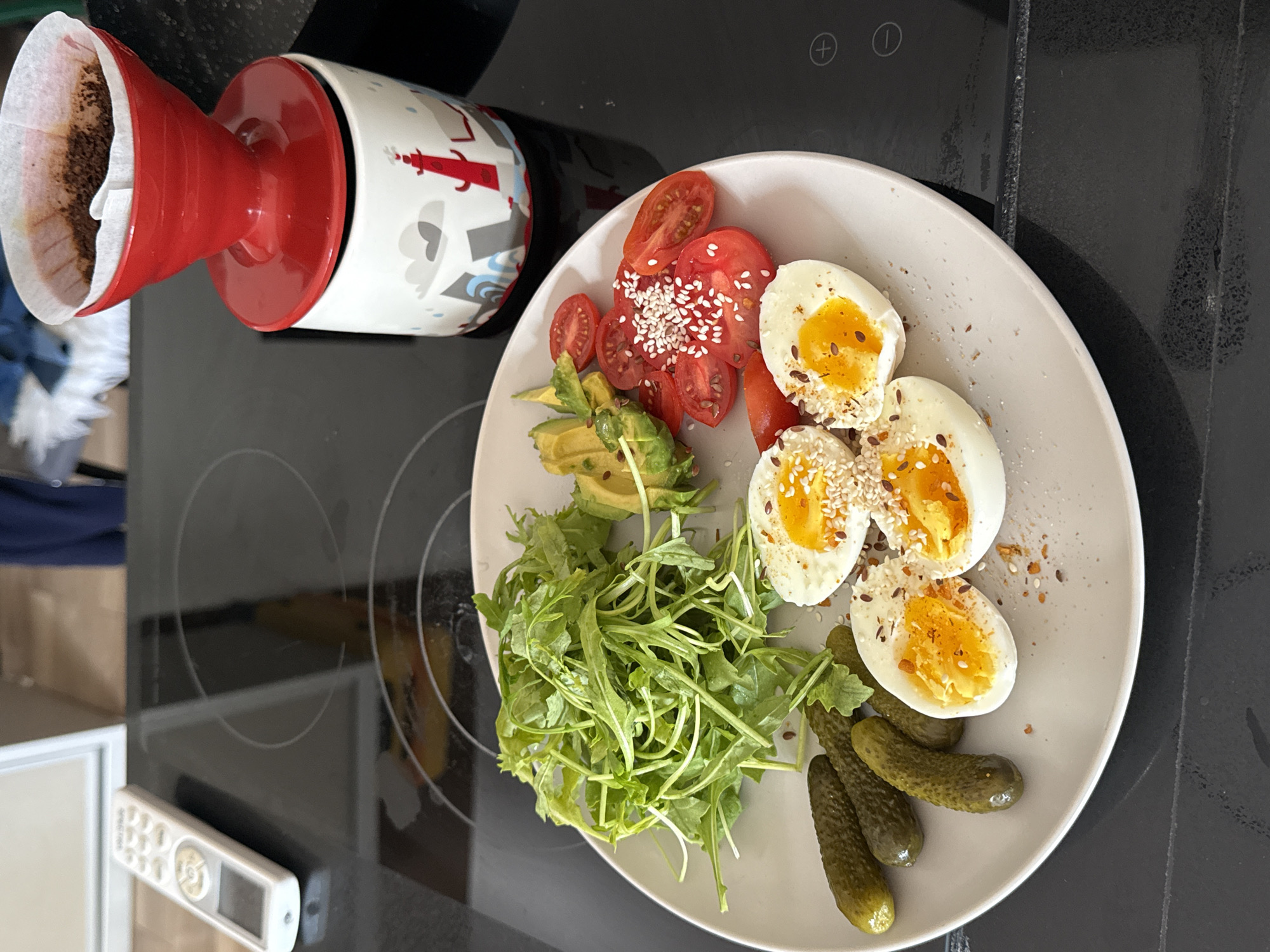 The scene presents a plate of food on a black stovetop, next to a coffee filter apparatus. The plate contains hard-boiled eggs cut in half, topped with seeds; sliced tomatoes with seeds; avocado slices; a pile of arugula; and several pickles. A coffee filter and a mug are visible next to the plate. The mug features a white background with cartoonish designs, including what appears to be a red lighthouse. The stovetop is a modern, flat electric type. The background lacks significant details to pinpoint a specific location in Tel Aviv, Israel.