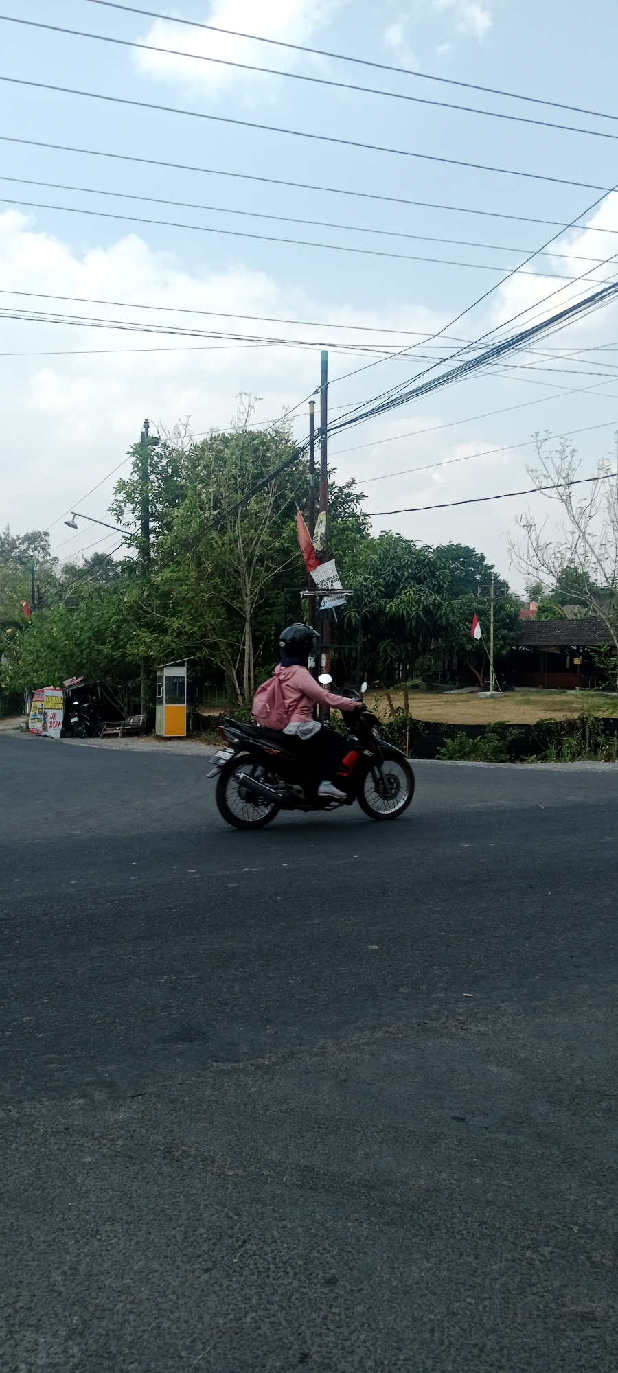 A person wearing a pink jacket and a black helmet is riding a black motorcycle on a paved road in Maguwoharjo, Indonesia. The motorcycle is in motion, with its wheels appearing to spin. The person is carrying a pink backpack on their back. In the background, there are trees, a small yellow and white booth, and power lines overhead. The sky is visible with scattered clouds. There are also some flags visible in the background, one of which appears to be an Indonesian flag. The scene appears to be on a street or road in a semi-urban or rural area.