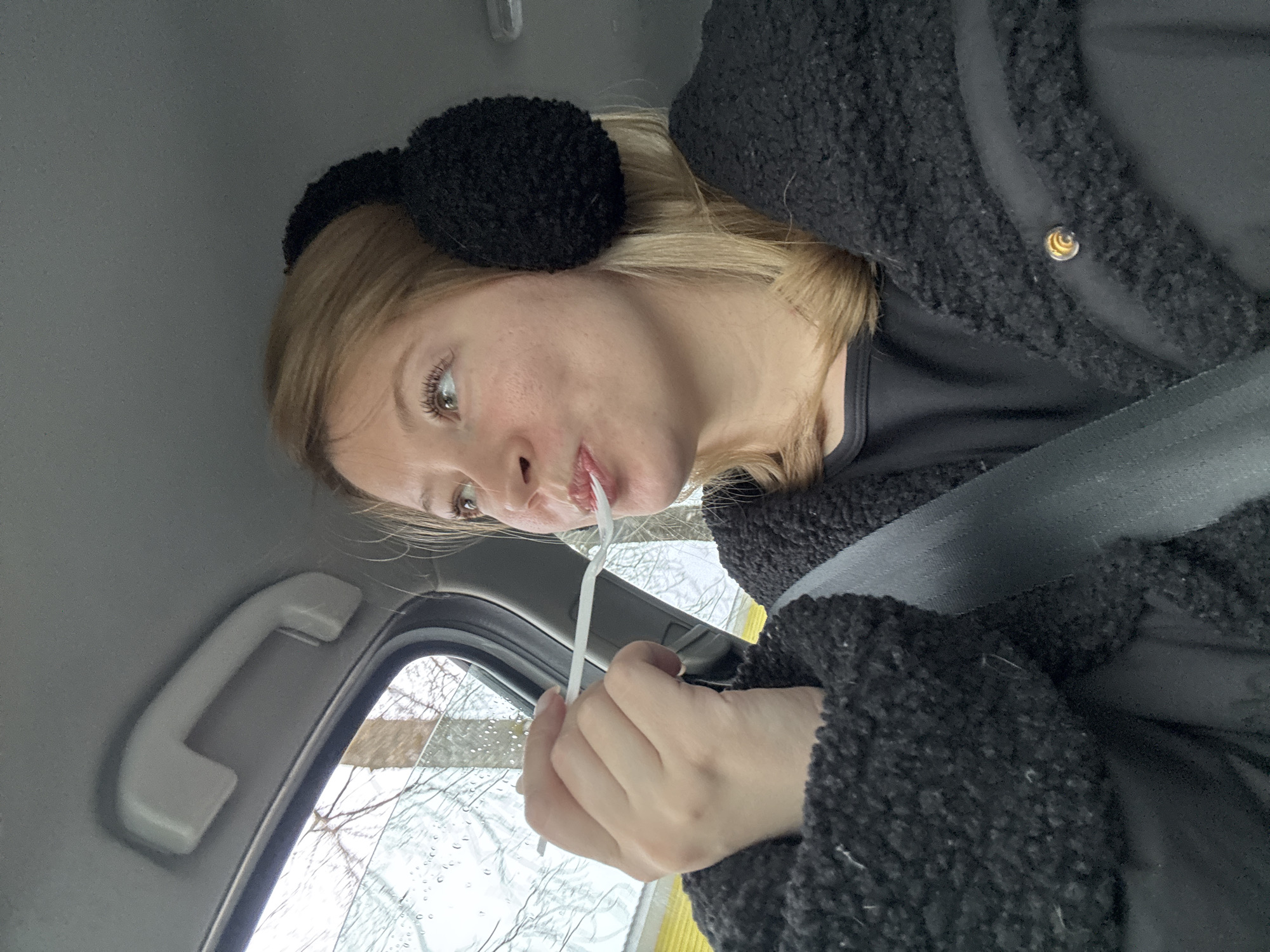 A woman is seated in a car, wearing black ear muffs and a black, fuzzy jacket. She has a seatbelt fastened across her chest. She is holding a white object, possibly a straw or strip of paper, to her mouth. The car window is visible, showing a blurred background of trees and what appears to be a building. Rain droplets are present on the window. Based on the prompt the location is Chaikovsky, Russia.