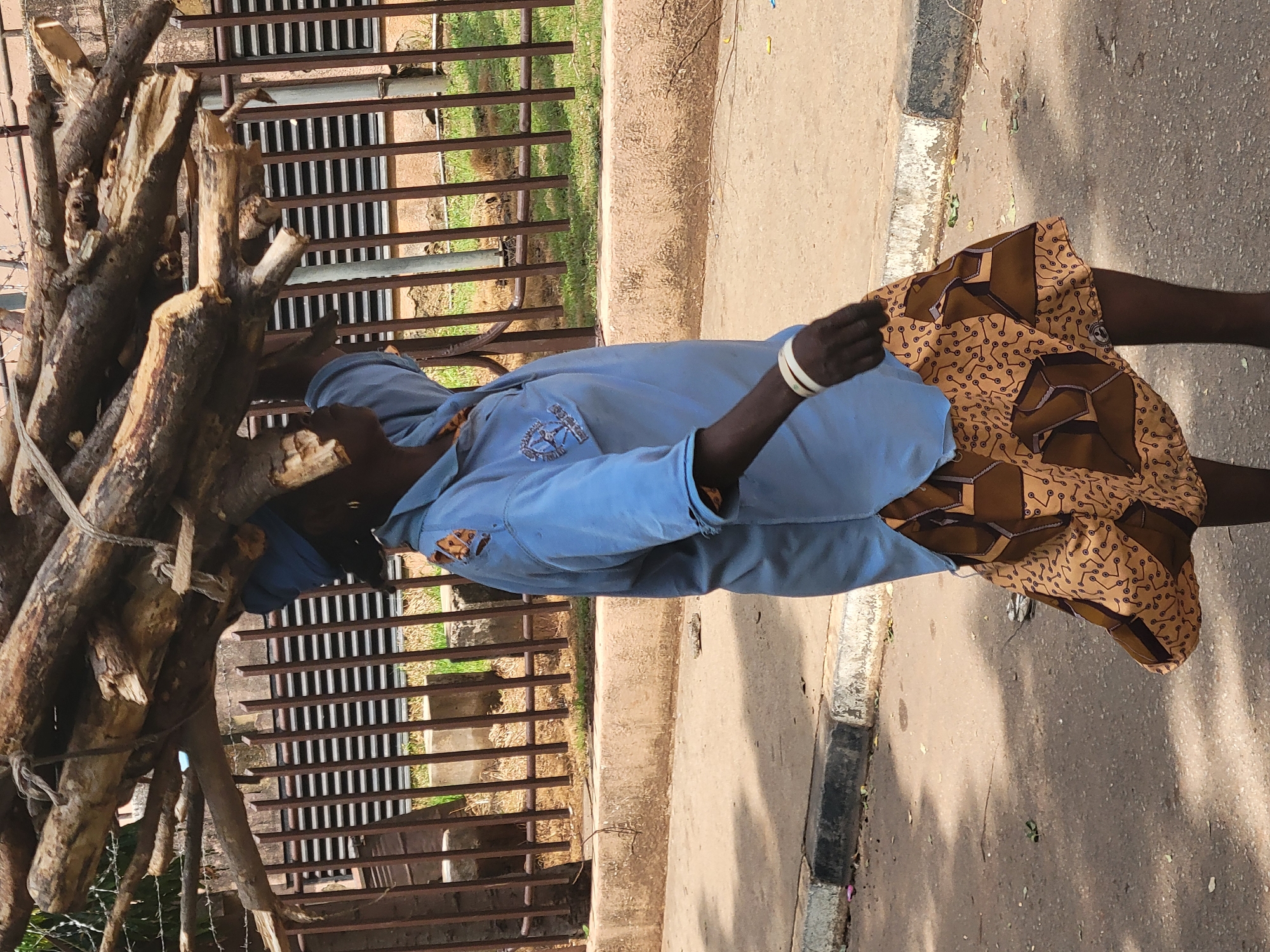 A woman in Abuja, Nigeria, is carrying a large bundle of firewood on her head. She is wearing a blue shirt, a brown and gold patterned skirt, and white bracelets on her arm. She is walking on a sidewalk next to a street. A brown metal fence is located to the left of the woman and the sidewalk has a grey curb.