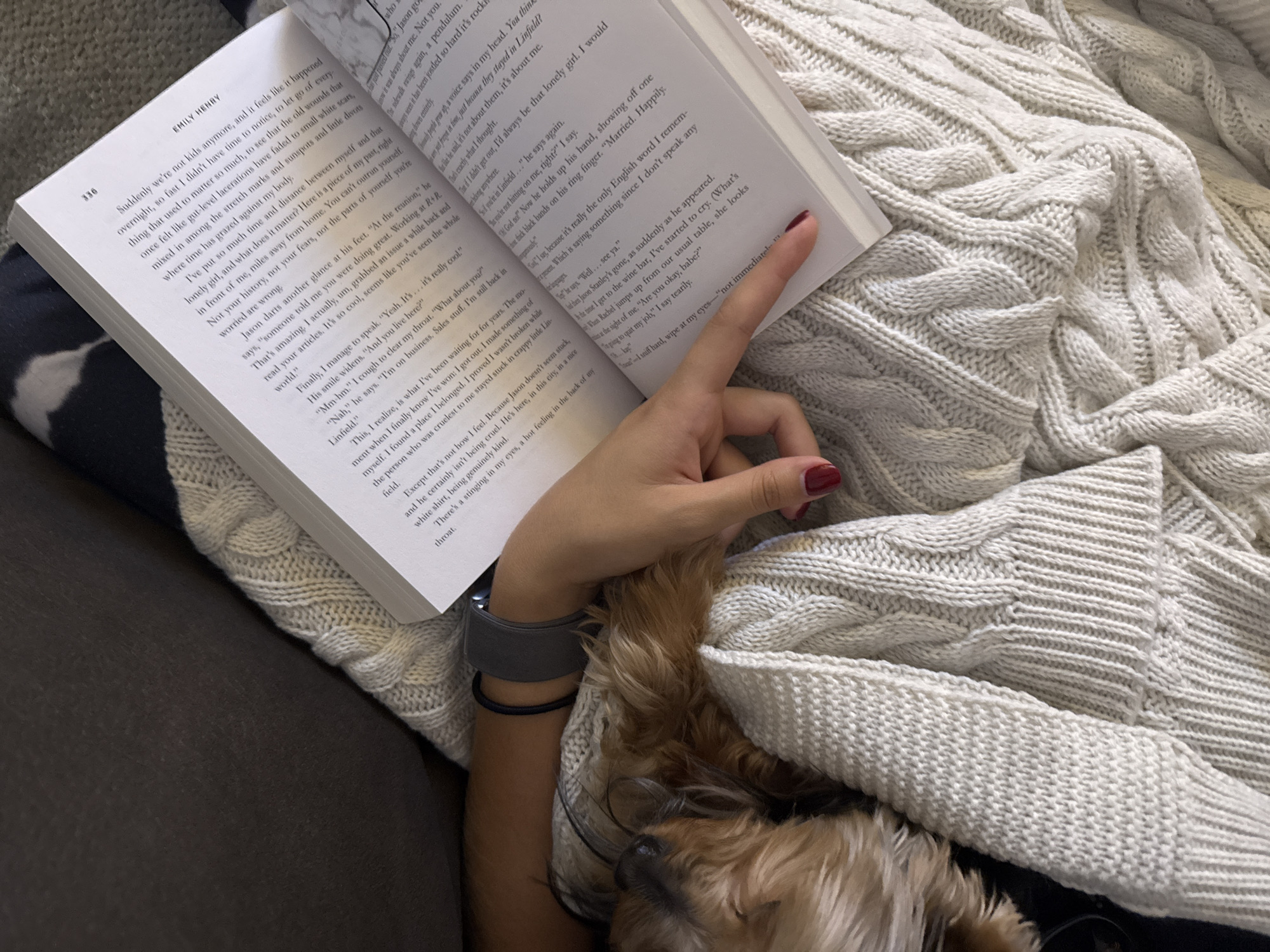 A person lies on a gray couch covered in a chunky knit blanket, reading the book "Happy Place" by Emily Henry. The person's left arm, adorned with a gray Apple Watch and black hair tie, rests on the blanket, their hand pointing to a passage on the book's right page. A small, tan dog is partially visible, nestled beside the person, under the blanket. The location is not San Bernardino Tlaxcalancingo, Mexico.