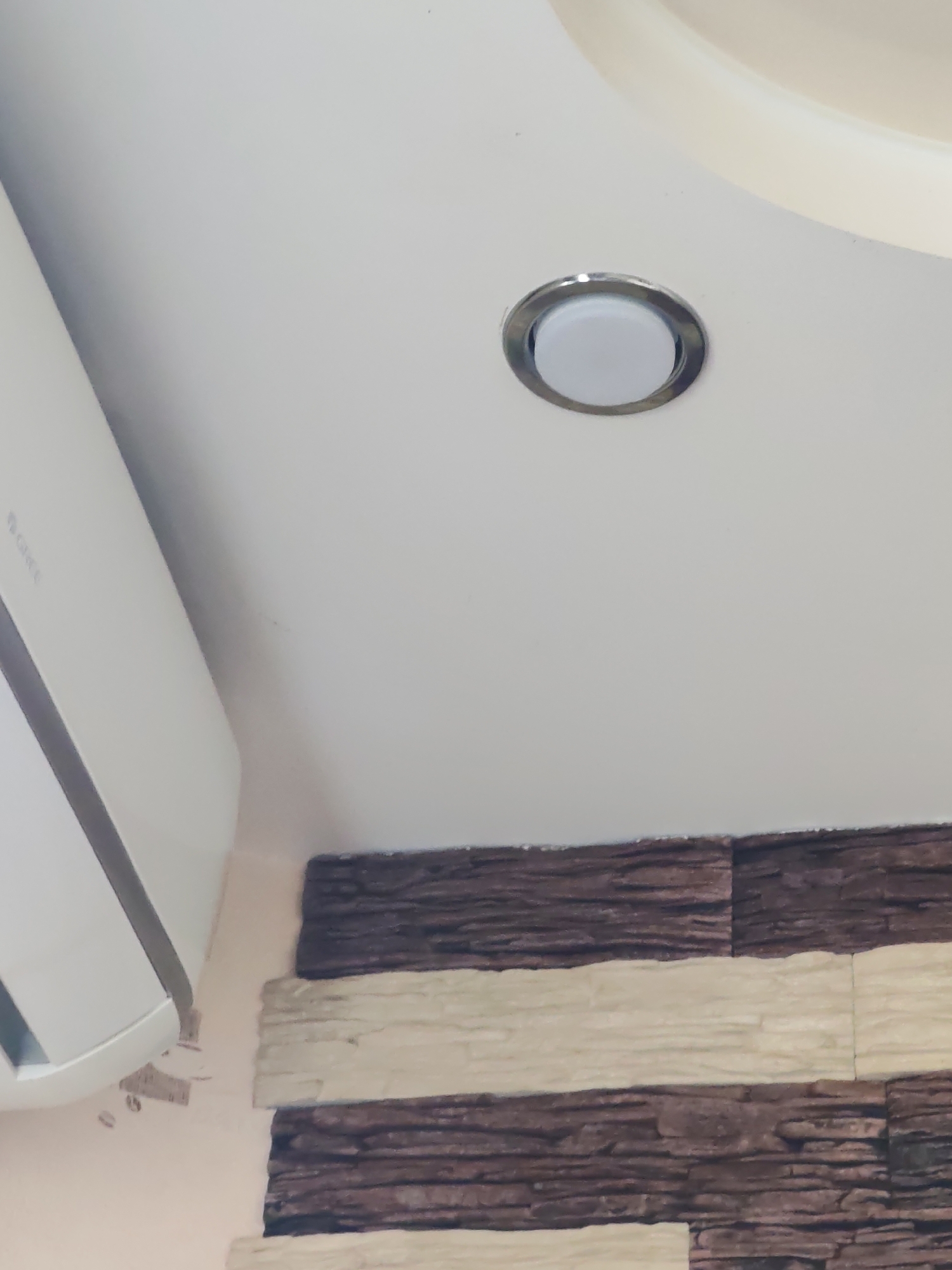 The image shows a partial view of an interior space. The elements in the frame are:
1. **Ceiling**: A portion of the ceiling is visible. It is white, with a slightly curved design. A recessed light fixture with a silver trim is present in the center of the frame.
2. **Air Conditioner**: The left side of the frame shows a white air conditioning unit.
3. **Wall**: The bottom part of the image displays a wall covered with what appears to be a textured stone veneer. The veneer is arranged in alternating horizontal bands of darker and lighter shades.
4. **Location**: The city and precise location within the city are unknown. No people, actions or interactions are apparent.