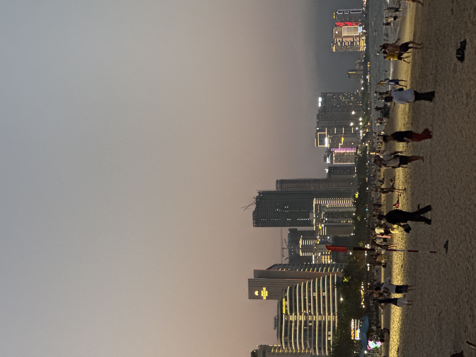 The image displays an evening scene on a sandy beach in Da Nang, Vietnam, facing an illuminated city skyline. The sky exhibits a muted gradient of grey and pale orange, indicating dusk or early night.
In the background, a dense cluster of high-rise buildings dominates the horizon. Many structures are brightly lit with architectural and decorative lights. Visible signage includes "NESTACELLIA" on a prominent, horizontally banded building and "FIVITOURANE" on a taller, more slender structure. Several construction cranes are discernible against the sky, indicating ongoing development.
The foreground and midground consist of a wide sandy beach populated by dozens of individuals. People are engaged in various activities, including walking, standing, and congregating in small groups. Some figures are visible near the water's edge on the right, where gentle waves are present. Patches of the sand are illuminated by artificial light sources originating from the beach or nearby urban area. A red flag or banner is affixed to a red and white striped pole, positioned centrally on the beach. A dark, amorphous object rests on the sand in the lower right quadrant. Tropical vegetation forms a visible line separating the beach from the urban infrastructure.