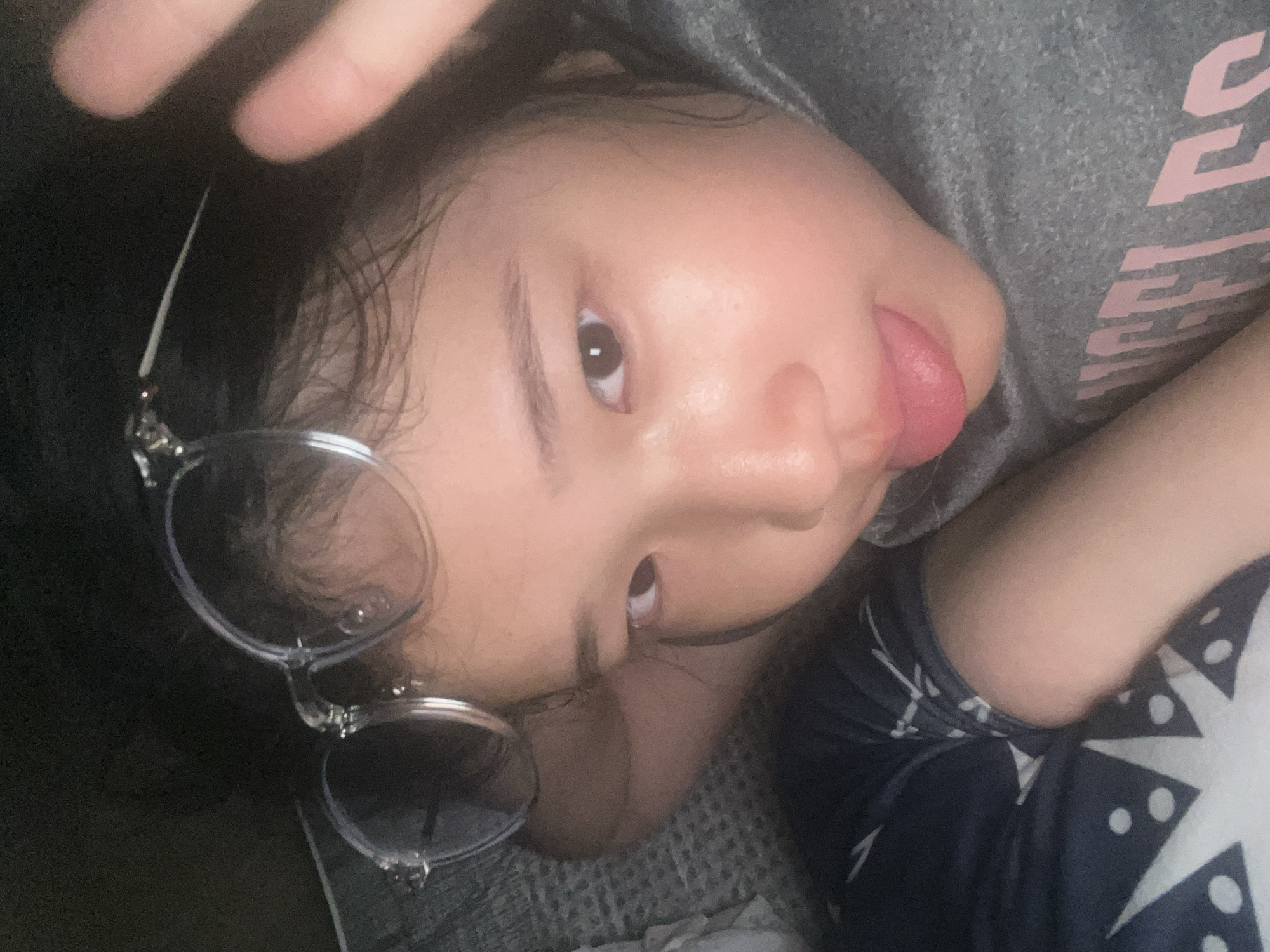 A person is lying on their back. The person is wearing glasses with round, silver frames. Their tongue is sticking out. They are wearing a gray t-shirt and a dark blue and white patterned garment. The location is Unknown, Unknown.