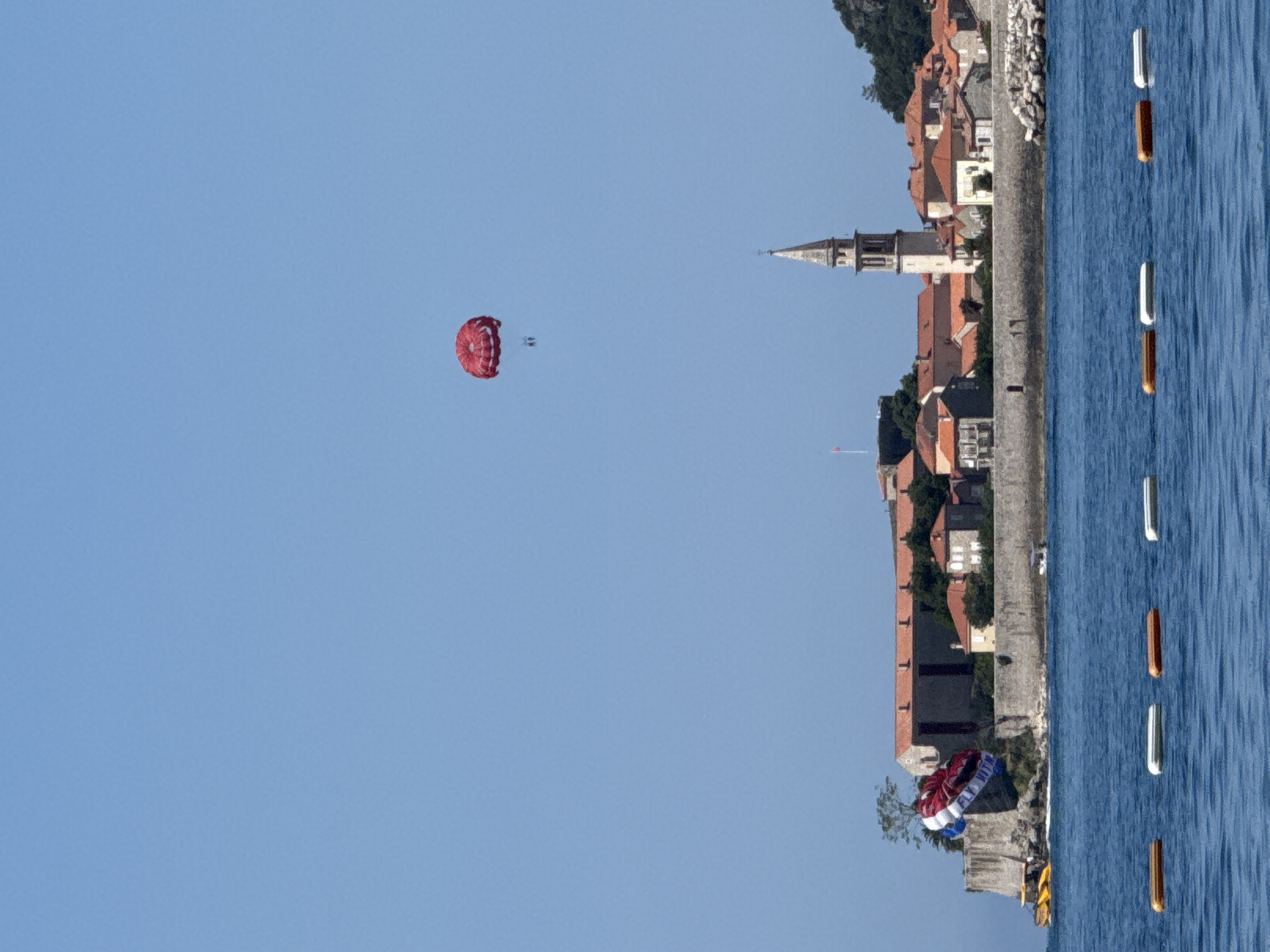The image displays a daytime coastal scene in Boreti, Montenegro, under a clear, light blue sky. In the foreground, blue water exhibits subtle ripples and contains a line of six rectangular buoys, alternating between orange and white. The midground features a town situated on a landmass extending into the water, characterized by stone walls along its perimeter and numerous buildings with terracotta-tiled roofs. A prominent, slender church tower with a spire rises above the town's structures on the right side. To the left, near the waterline, a yellow watercraft is visible alongside a partially deflated multi-colored parachute (red, white, and blue), which displays the partial text "FLY WIT". High in the upper-right portion of the sky, a red parasail is airborne, with two small human figures suspended directly beneath it.