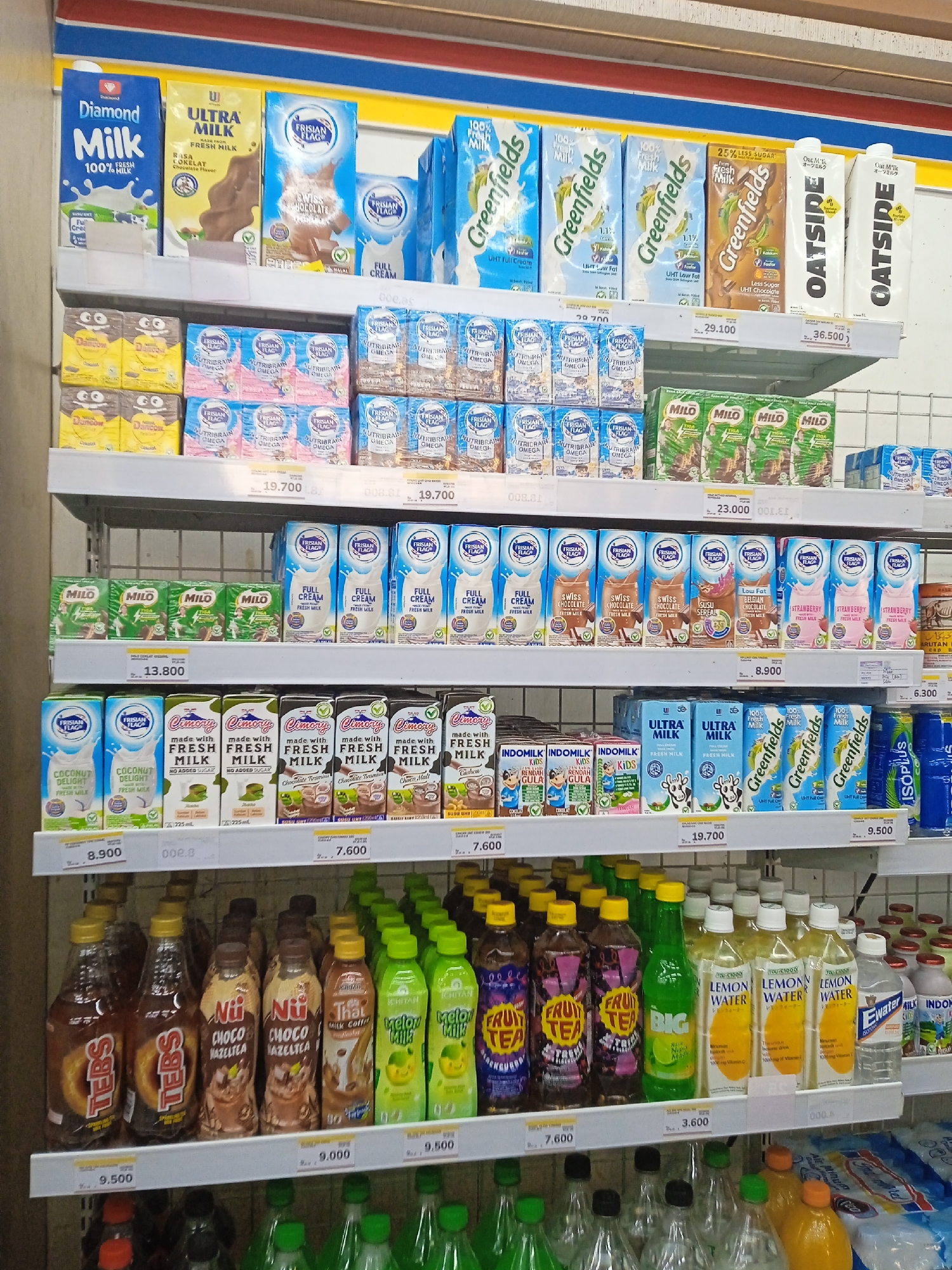 This is a medium shot, eye-level photograph of a retail store shelf filled with various dairy and beverage products. The products are neatly arranged in their packaging, primarily cartons and bottles, on white shelves. The lighting appears to be artificial and bright, typical of a convenience store.
On the top shelf, from left to right, are cartons of "Diamond Milk," "ULTRA MILK," and several cartons of "Frisian Flag" branded milk, including "Full Cream" and "Chocolate" flavors. Further to the right are "Greenfields" branded milk cartons, "OATSIDE" oat milk cartons, and a white carton with yellow and black text.
The second shelf from the top displays more cartons, including what appear to be "Dancow" products, "Frisian Flag Nutribrain Omega," and "Milo" products.
The third shelf features a variety of milk cartons. On the left are "Frisian Flag Coconut Delight" and "Frisian Flag Full Cream" cartons. In the center are "Cimory made with Fresh Milk" cartons in various flavors like "Chocolate Vanilla," "Chocolate Malt," and "Cashew." To the right are more "Frisian Flag" cartons labeled "Swiss Chocolate," "Susu Ceres," "Belgian Chocolate," and "Strawberry."
The fourth shelf contains "Frisian Flag" milk cartons on the left, followed by "Indomilk Kids" cartons, and then "ULTRA MILK" cartons. To the right are more "Greenfields" milk cartons and a blue bottle of "Iso Plus" electrolyte drink.
The bottom shelf prominently displays a row of bottled drinks. On the left are bottles of "TEBS" soda. Next are several bottles of "Nii Choco Hazelnut" and "Nii Choco Matcha" flavored drinks. Further to the right are bottles labeled "That Milk Coffee," "Ichitan Big," "Ichitan Fruit Tea Strawberry," and "Ichitan Melon Milk." Some bottles of "Lemon Water" and "Ewater" are also visible, along with some dairy cartons on the far right.
The background shows a tiled wall, typical of a store environment, with some shelving hooks and possibly other merchandise out of focus. The price tags are visible below most of the products on the shelves. The overall impression is an organized retail display of beverages. The image was taken in Kaliwungu, Indonesia, likely within a supermarket or minimarket. No people are visible in the image.