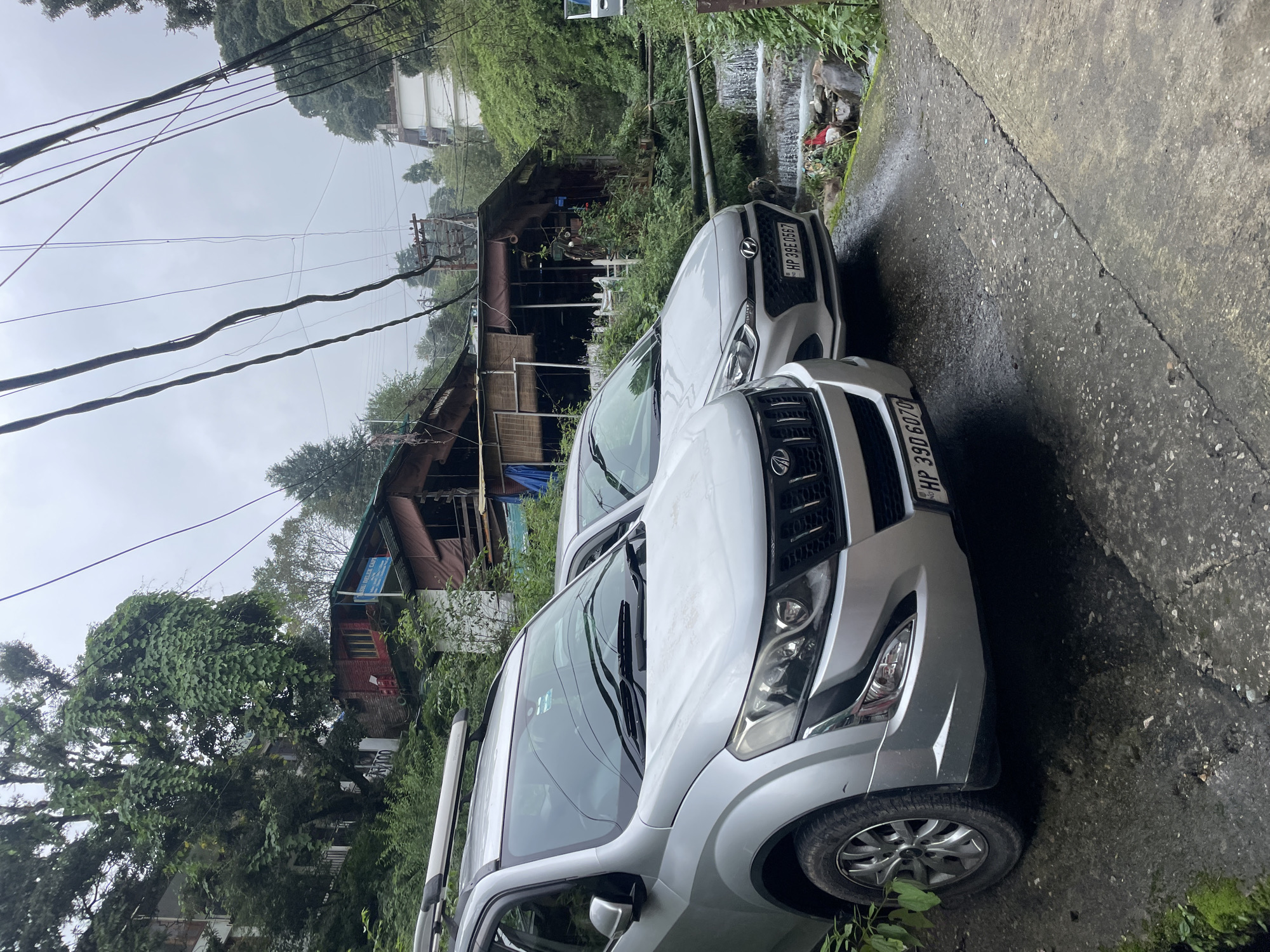 Two silver SUVs are parked on a wet, paved road in Bhagsu, India. The SUV in the foreground is a Mahindra XUV500 with license plate "HP 39D 6070". The SUV behind it is a Hyundai i20 with license plate "HP 39E 0567". In the background, there are multi-story buildings with sloped roofs, dense green foliage, and utility wires crisscrossing overhead. A small waterfall cascades into a rocky stream to the right of the vehicles. The sky is overcast.