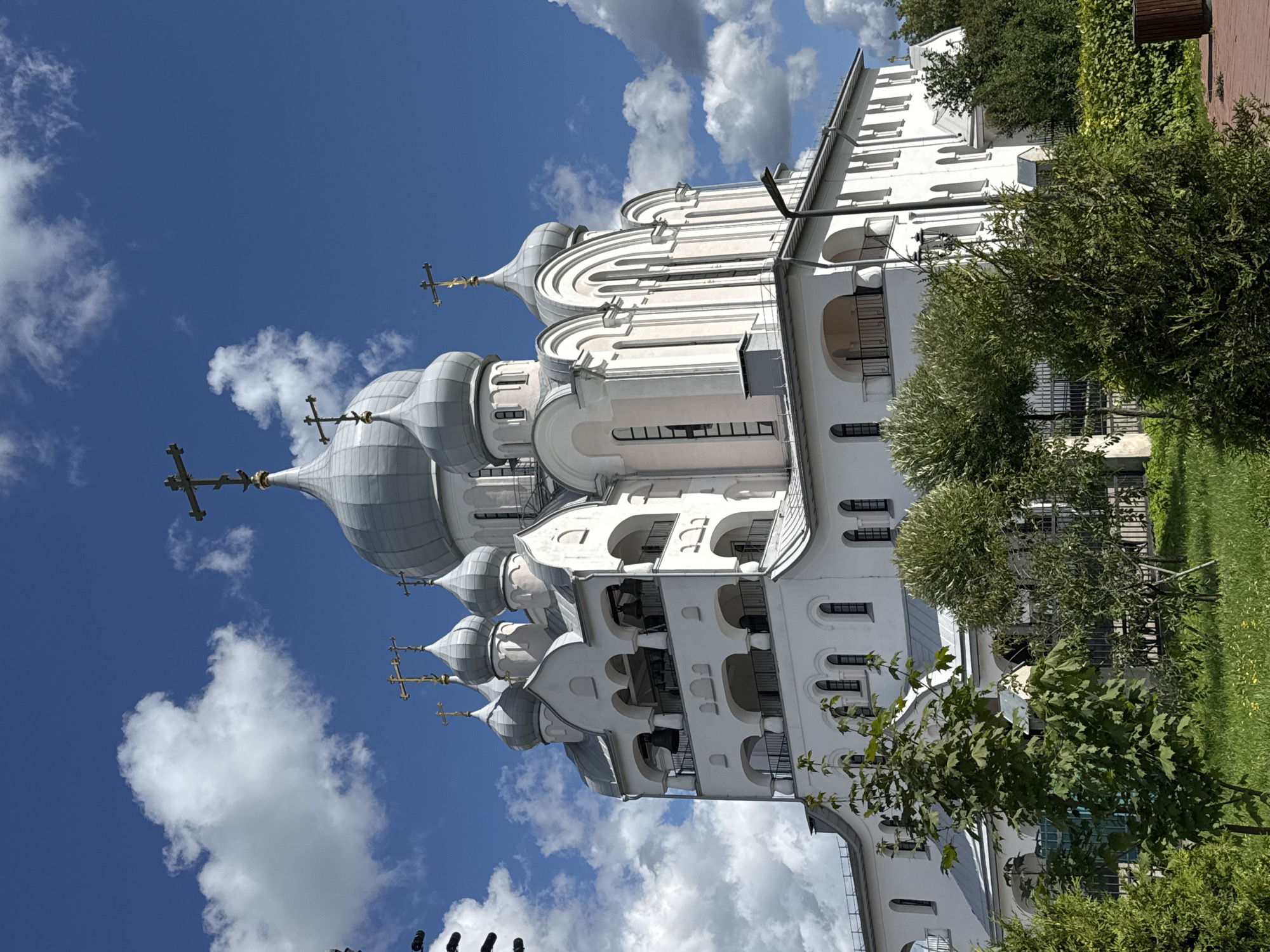 A grand, white Orthodox church with multiple silver domes topped with golden crosses stands against a vibrant blue sky dotted with white cumulus clouds. The church features a bell tower with arched openings and several balconies. Lush green trees and grass surround the building, with a paved walkway visible in the foreground. The image was taken on a sunny day, likely during daylight hours. The architecture suggests a location in Saint Petersburg, Russia, known for its numerous historical and religious structures. There are no people or animals visible in the image.