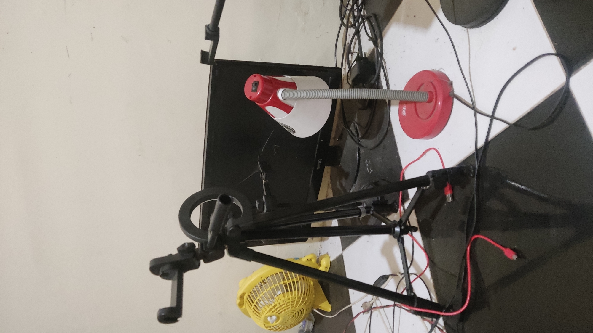 The image depicts a cluttered indoor scene, likely a home office or a creative space, with various electronic devices and equipment. Dominating the foreground is a black tripod with a phone mount and a ring light attached. Red USB cables are visible, connecting to the tripod and running across the floor.
To the left of the tripod, a bright yellow electric fan is positioned. Behind the tripod, a black computer monitor with a "BENQ" logo is present. In front of the monitor and to the right of the tripod, a red and white gooseneck desk lamp with an on/off switch on its shade is situated on the floor.
The floor has a checkered pattern of black and white tiles. The background wall is a neutral, light color. The overall impression is one of a functional but somewhat disorganized setup for content creation or personal computer use. The location of Ngadiharjo, Indonesia, is an extraneous detail not deducible from the visual evidence.