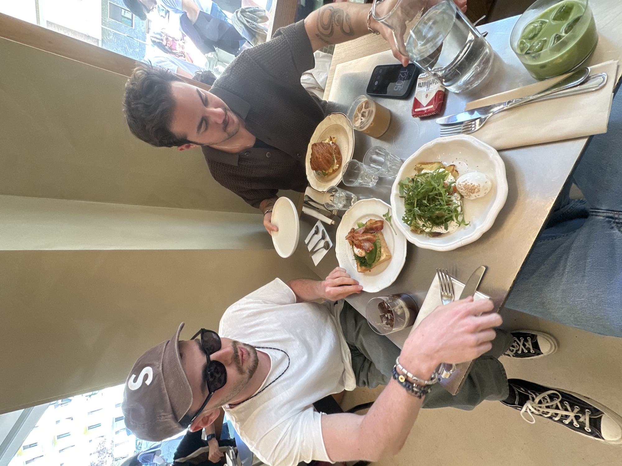 Two men are at a table with food and drinks. One man is pouring water from a glass pitcher. On the table are plates with food including eggs and arugula, a sandwich, and drinks. There is a pack of Marlboro cigarettes and a cell phone on the table. One man is wearing a hat with a white "S" and sunglasses. The other man has tattoos on his arm. The location may be a restaurant in Milan, Italy, based on the architecture seen through the window in the background.