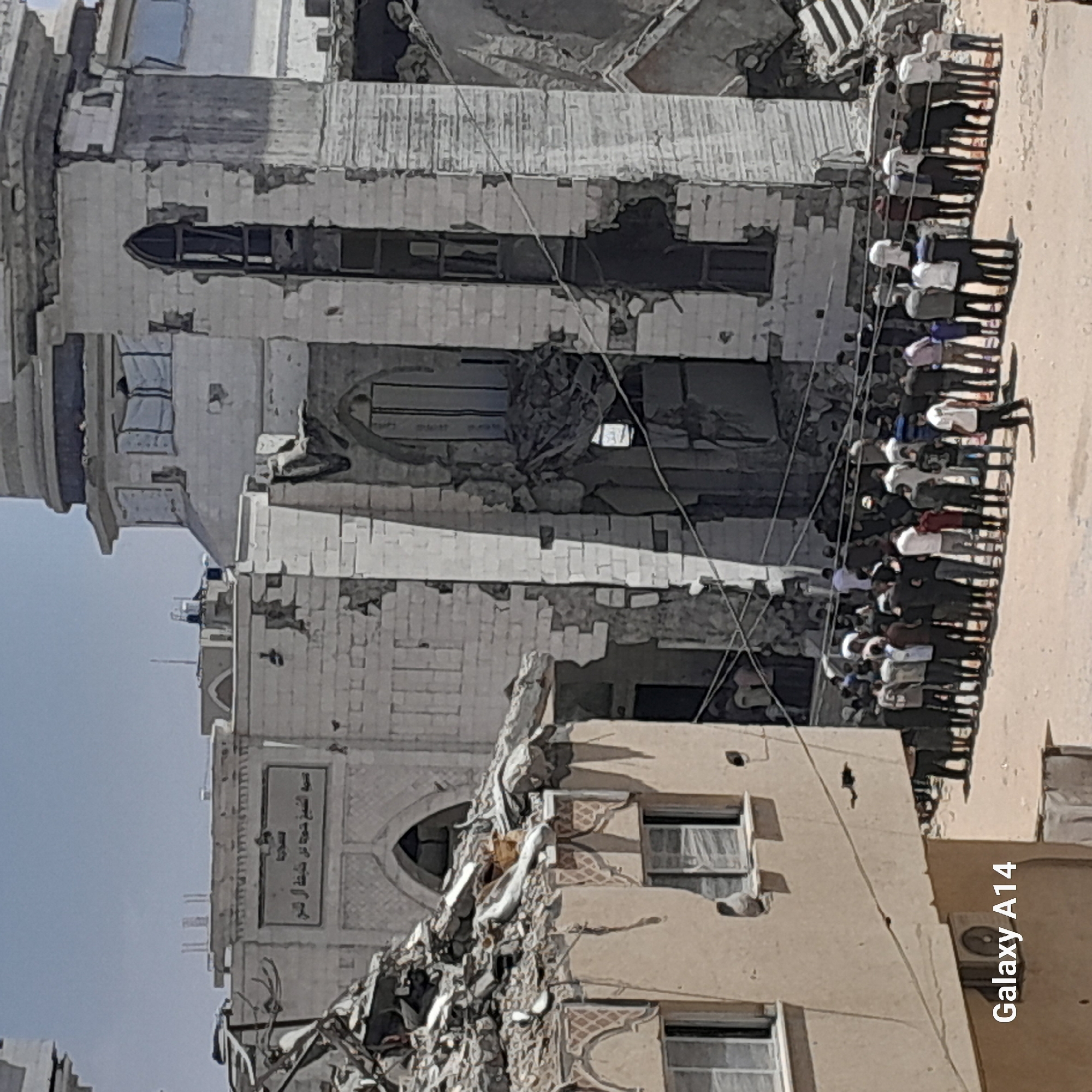 A group of people are gathered in the street in front of damaged buildings, one of which appears to be a mosque, in Khan Yunis, Palestinian Territories. The buildings show signs of destruction and rubble. Many of the people are standing in a line, facing the damaged buildings. The sky is clear and the sun is shining. The image is a photograph taken from a slightly elevated position.