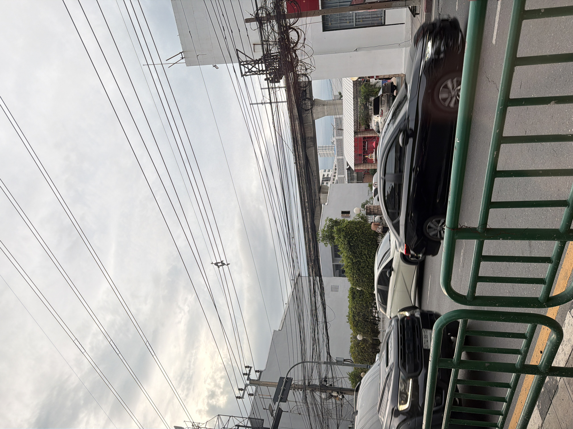 This is a street-level view of an urban environment in Bangkok, Thailand, under an overcast sky. The foreground features a green metal fence with yellow markings on the asphalt behind it. A black SUV is partially visible on the left, and a black sedan is in motion on the right, blurred by its speed. In the middle ground, several cars are on the road, including a white sedan. Buildings with white facades and green bushes line the street. Overhead, a complex network of electrical wires crisscrosses the sky, some of which appear to be attached to utility poles. A concrete elevated structure, likely for public transportation, runs horizontally across the upper portion of the image. The scene depicts a typical busy street in Bangkok with traffic and extensive electrical infrastructure.