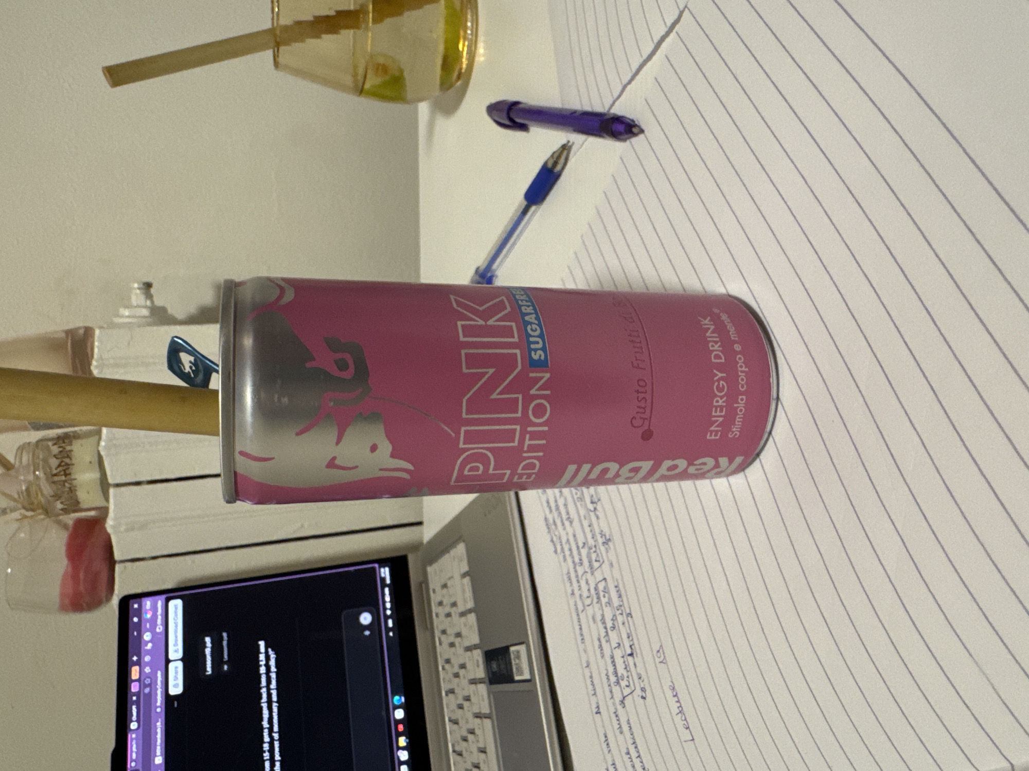 The image shows a desk setting, possibly in Milan, Italy, indicated by the Italian writing on the Red Bull "Pink Edition" can. The desk contains a laptop displaying a PDF titled "Lesson19.pdf," a lined notebook with handwritten notes ("Lecture 19"), a blue pen with a purple cap, and a glass with a yellow liquid and a wooden stick. A decorative glass bottle and a jar with red liquid are in the background.
