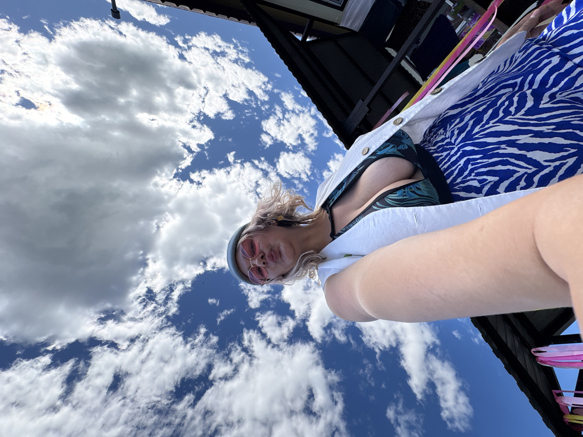 A low-angle, wide shot captures a woman in a light-colored jacket, a dark patterned swimsuit, and a blue and white zebra-print skirt, posing outdoors against a bright blue sky filled with fluffy white clouds. The woman, with blonde hair and wearing pink-rimmed sunglasses and a light blue hat, puckers her lips as if for a kiss. The background features the dark, slanted roof of a building with visible trim, and decorative colorful ribbons are attached to the roofline. The image does not provide any specific visual cues to indicate a location in Birobidzhan, Russia.