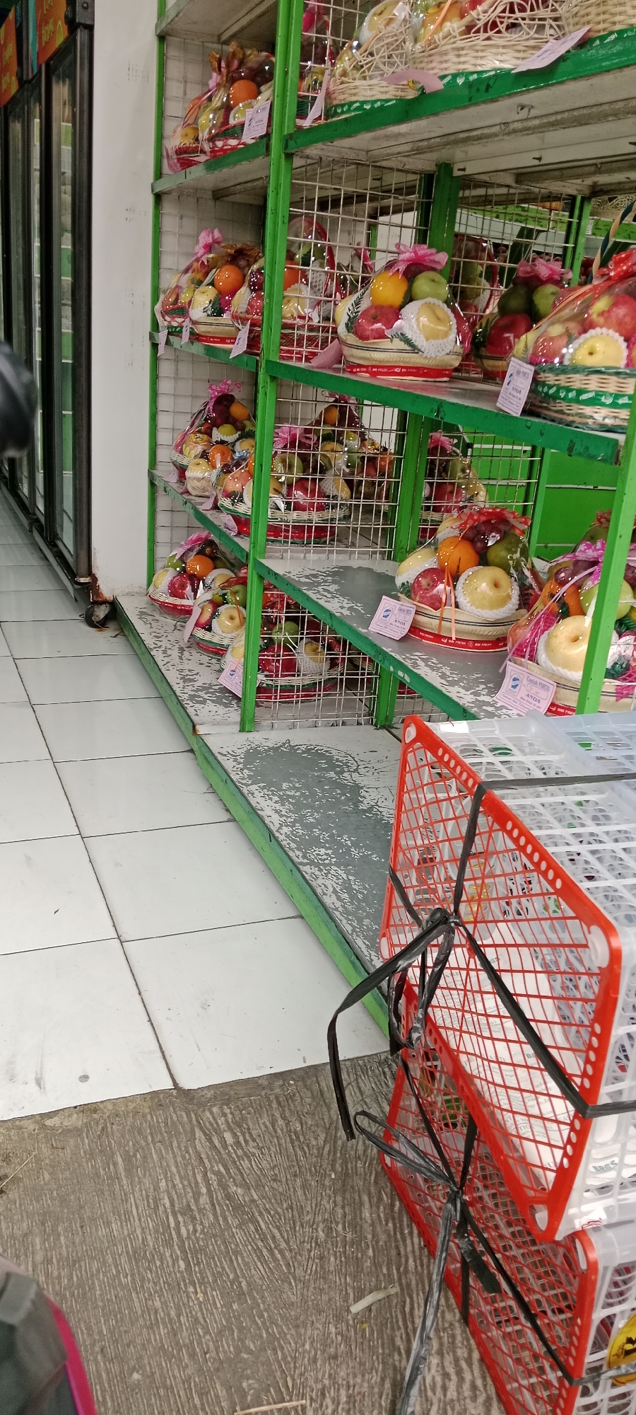 The image shows a retail environment, likely a market or supermarket, with shelves stocked with fruit baskets. The shelves are constructed from green metal frames with wire mesh shelving. The fruit baskets are individually wrapped in clear plastic and decorated with pink ribbon. Visible fruits include apples, oranges, grapes, and pears. Some baskets have labels attached. In the foreground, two stacked plastic crates, one red and one white, are visible, secured with black plastic ties. The floor is tiled with white square tiles, and a section of textured grey concrete is also present. The background features a display cooler with glass doors. The overall scene suggests a commercial setting where produce is being sold.