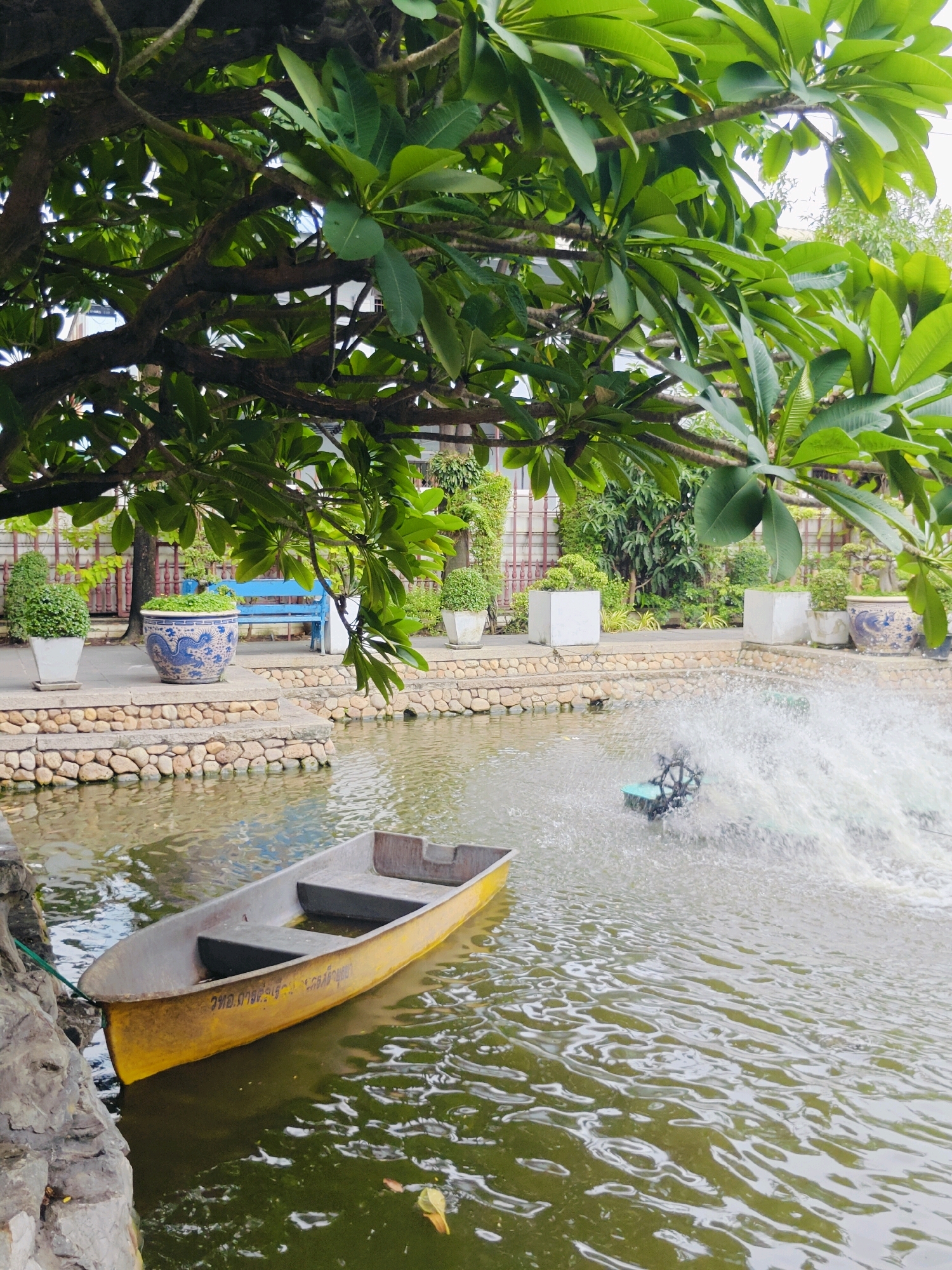 This image depicts a tranquil canal-side scene in Bangkok, Thailand. A weathered, yellow rowboat is moored near a stone embankment on the left. In the middle ground, a water aerator churns up white spray, its black paddle wheel visible. The canal is bordered by a stone pathway and a low wall constructed from rounded river stones. Behind the wall, a blue bench sits on a paved area, and various potted plants and decorative urns are placed along the walkway. Lush green foliage from a large tree drapes over the scene from the upper left. The overall impression is one of a peaceful outdoor setting, likely a park or garden area, with elements of both natural beauty and urban design.