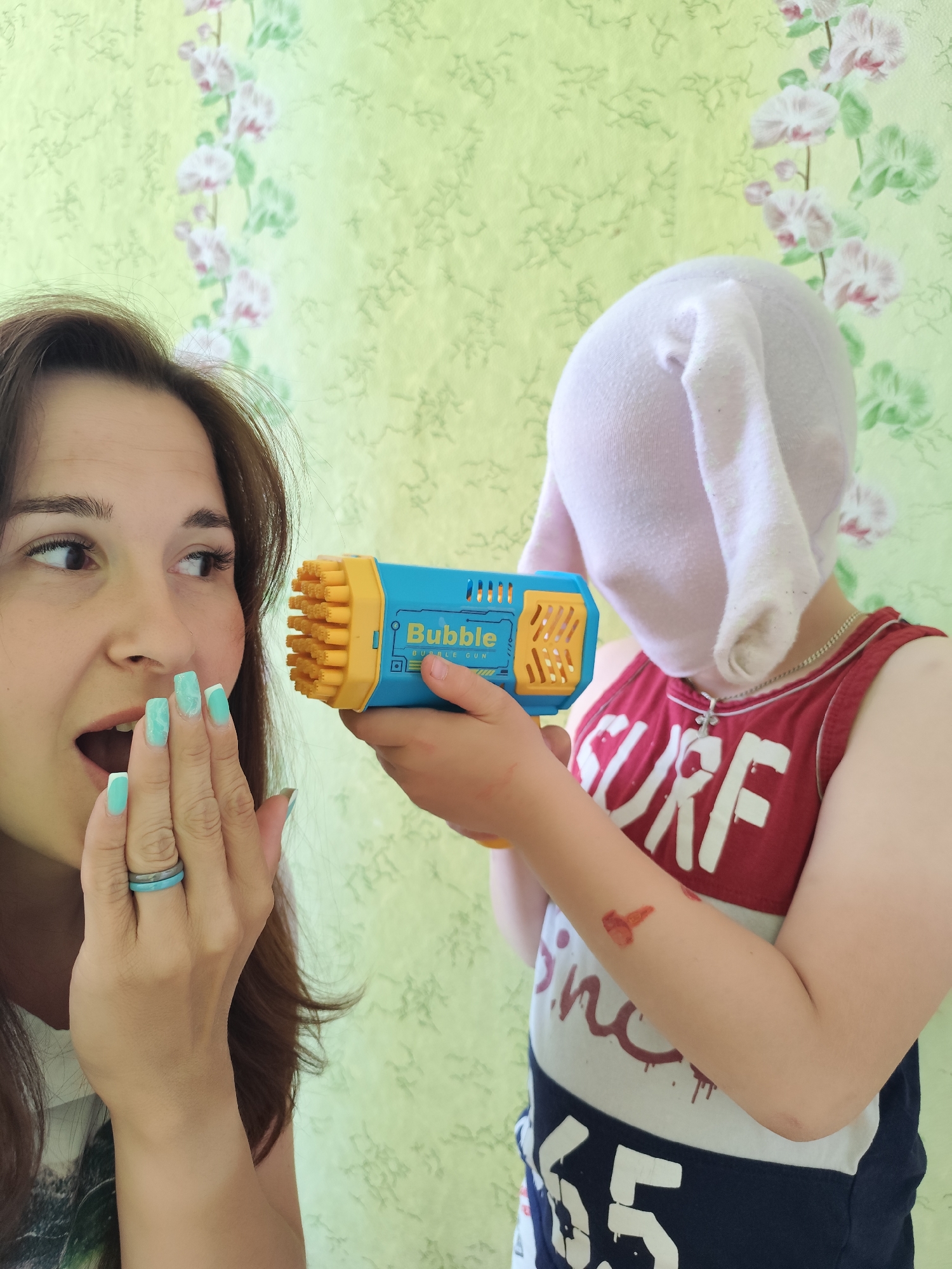 A woman with brown hair and turquoise nails appears surprised, with her mouth open and hand near her face. A child wearing a white head covering that resembles rabbit ears is holding a blue and yellow "Bubble Gun" towards her. The child is wearing a red, white, and blue "Surf" tank top and a silver necklace. The background features a light green wallpaper with a floral pattern. The location is in the city of Фоки, Russia.