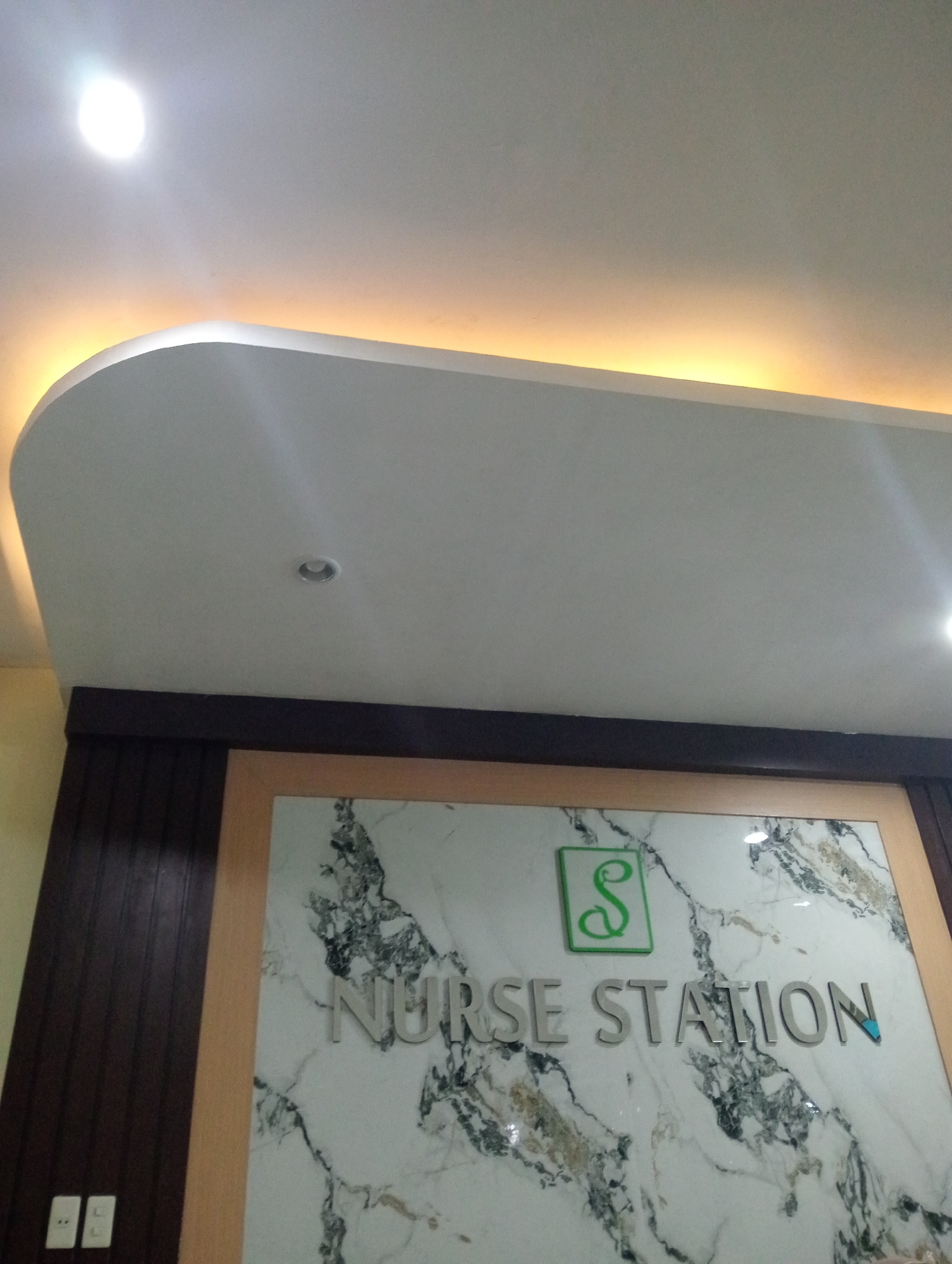 The image shows a close-up of a sign that reads "NURSE STATION" in silver lettering against a marble-patterned background. Above the text, there is a green square logo with a stylized "S" inside. The sign is framed by wooden trim. To the left of the sign, there is a dark brown paneled wall with two white electrical outlets. The ceiling above is white with recessed lighting and a soft yellow glow emanating from a cove light. The overall impression is of an indoor setting, likely a healthcare facility, given the signage. The image appears to be taken at the Nurse Station of RSUD Pringsewu in Pringsewu, Lampung, Indonesia.