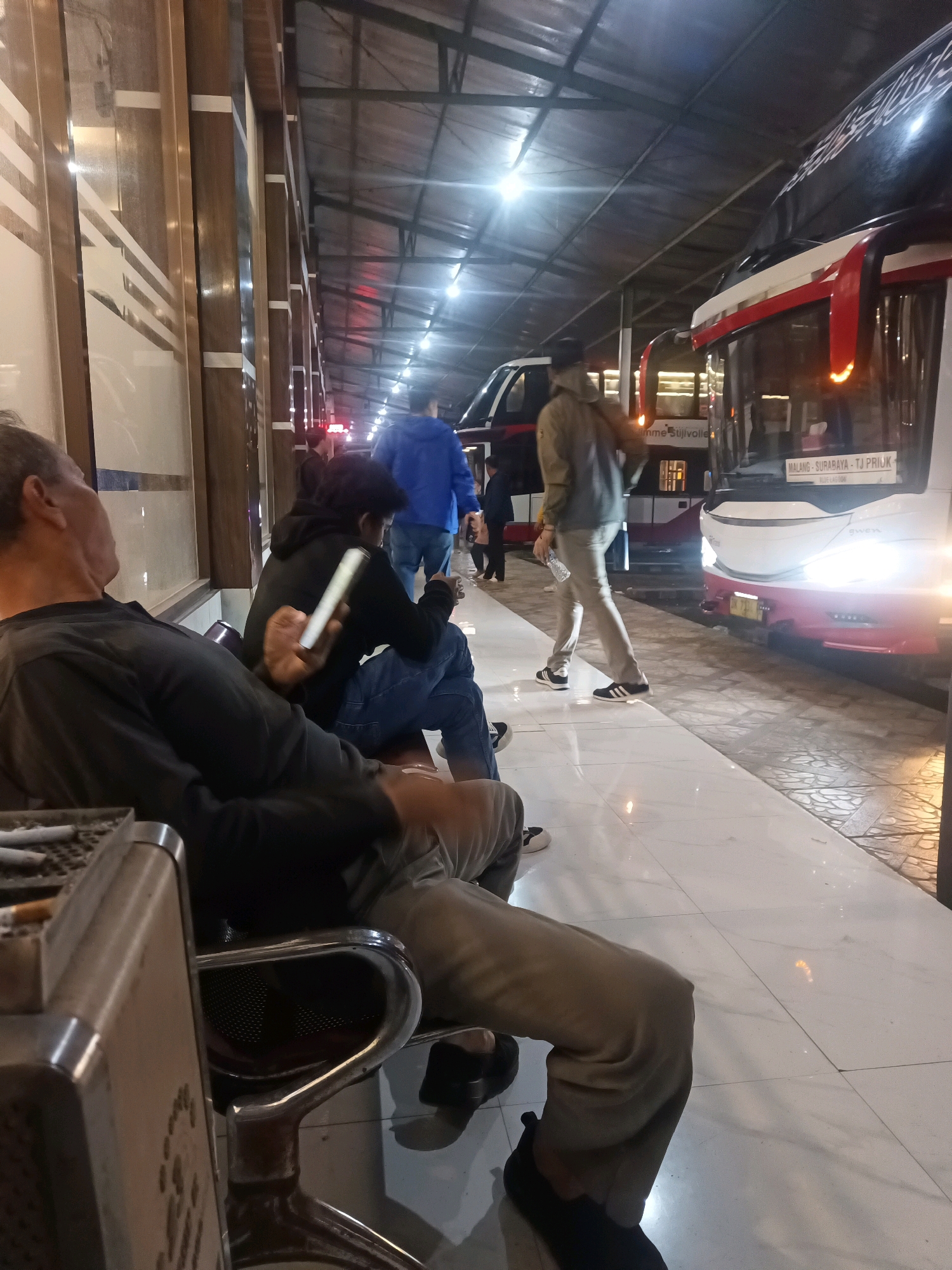 At a bus station in Cikawung, Indonesia, several individuals are waiting. In the foreground, a man with graying hair is seated, wearing a black long-sleeved shirt and light-colored pants. He is holding a white smartphone. To his right, another person in a black hoodie and blue jeans is also seated, looking at a white smartphone. Behind them, a person in a blue jacket walks away, followed by a person in a light brown jacket who is holding a water bottle. Two buses are parked at the station. The bus on the right is white with red accents and displays "MALANG - SURABAYA - TJ. PRIUK" on its front. The bus on the left has a dark exterior with red trim. The environment includes a covered waiting area with tiled flooring and overhead lighting. A metal ashtray with several cigarettes is visible in the lower left corner.