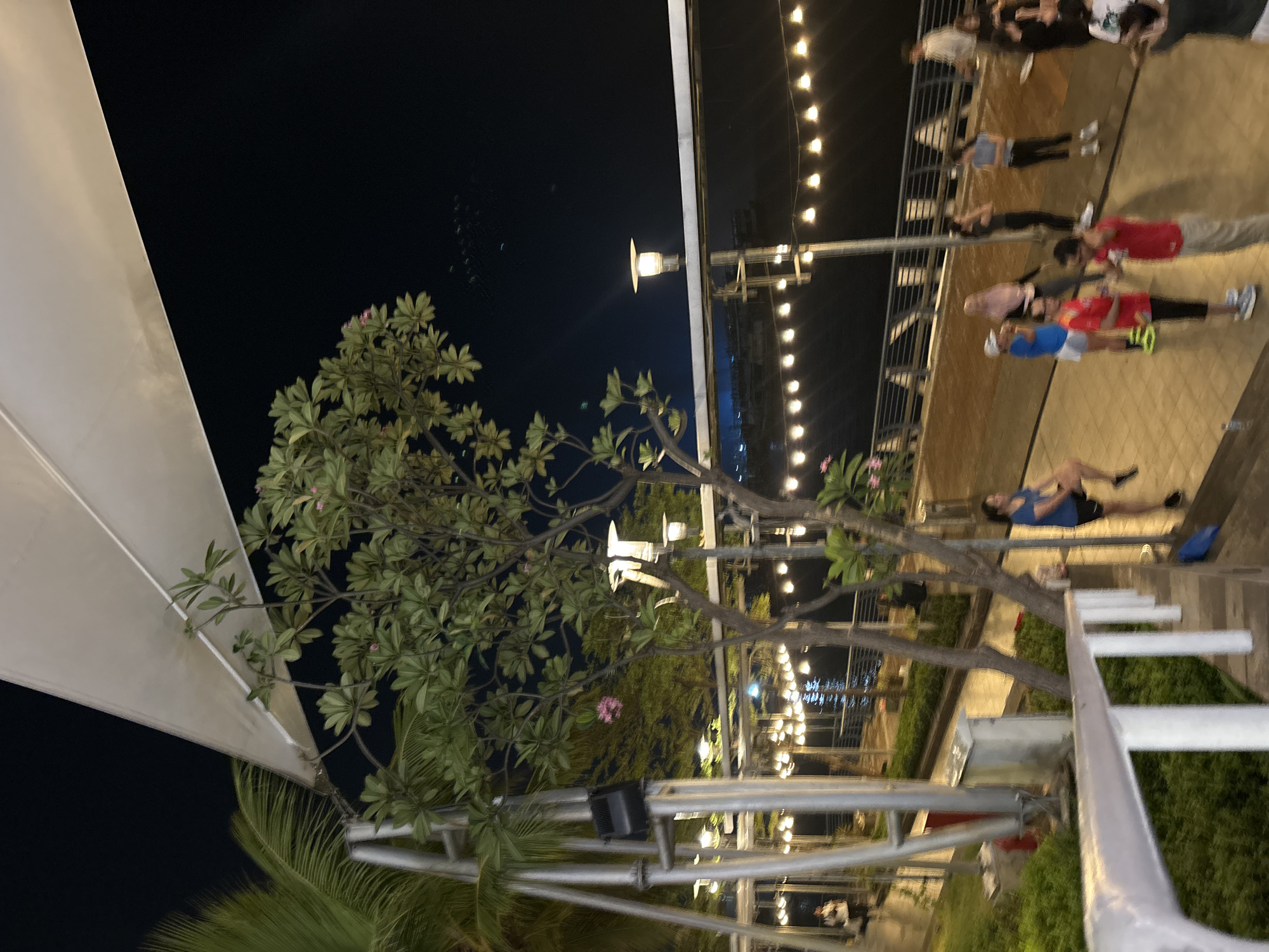 A nighttime scene unfolds at Ancol Beach City, North Jakarta, Indonesia, featuring an outdoor promenade along the waterfront. A large white canopy stretches across the upper portion of the frame, providing shade and structure. Lush green trees with delicate pink flowers are scattered throughout the scene, some with palm fronds visible. The promenade is illuminated by a string of warm, yellow lights that cast a gentle glow, and several streetlights are also present.
A group of people are engaged in various activities. Some appear to be exercising or stretching, while others are standing and conversing. A woman in a blue top and black shorts is captured mid-stride, her leg raised. Another woman in a light blue headscarf and white shorts stands near a man in a red t-shirt and black leggings. A group of women in athletic wear are also visible, some with hands on their hips or engaged in conversation. The water of the bay is visible in the background, reflecting the lights of the city. The overall atmosphere suggests a casual evening gathering or fitness activity.