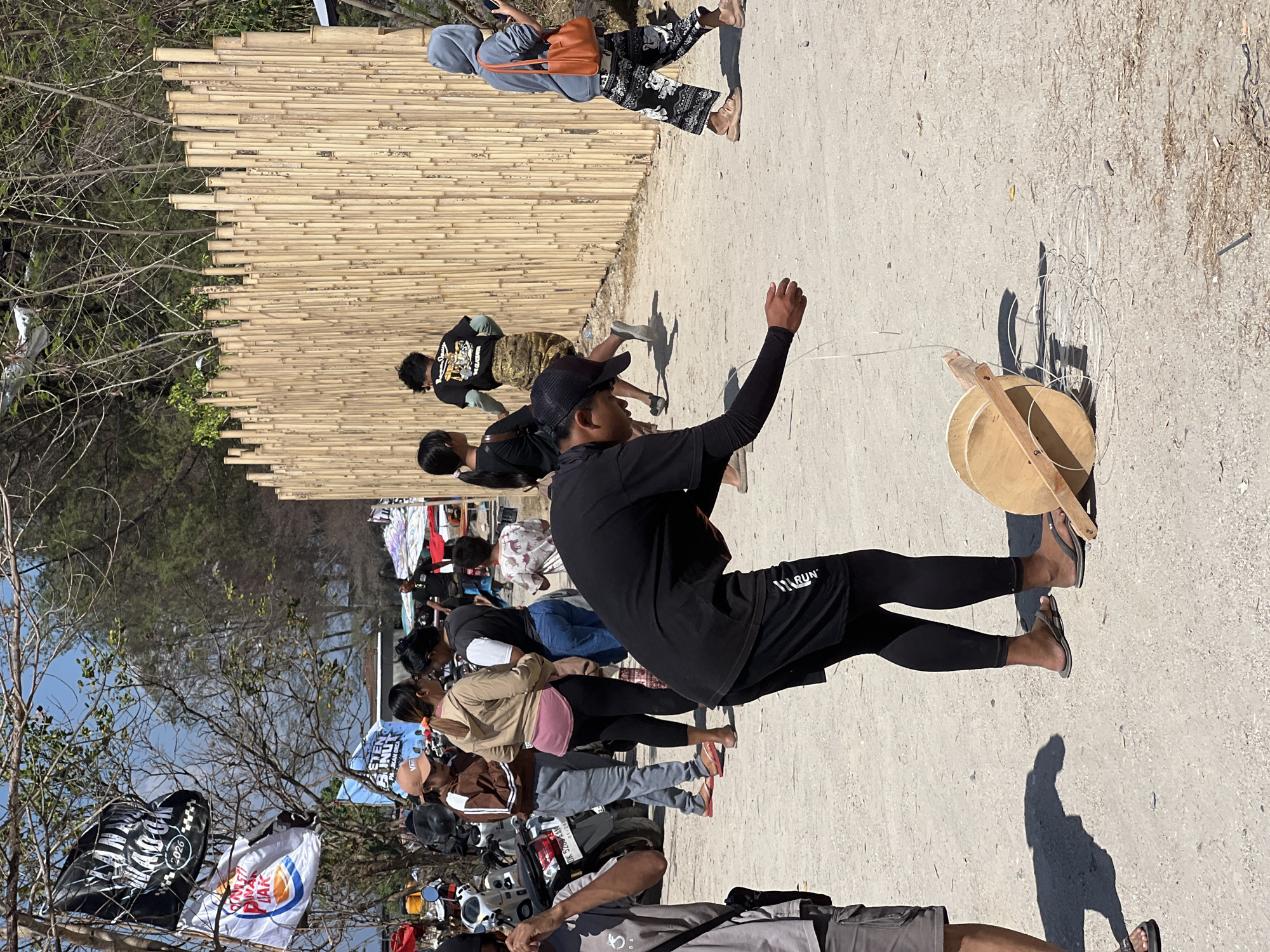 The image depicts a dusty outdoor scene, likely a public area in Denpasar, Indonesia, possibly near a beach given the presence of trees and a bamboo fence. In the foreground, a person wearing black athletic wear and a black cap is crouched down, manipulating a large wooden spool of white string. The spool rests on the ground, and the string is partially unraveled around it.
Several other individuals are visible in the midground and background. To the left, a group of people stand near several parked motorcycles, with one person wearing a brown hoodie and another a light-colored hooded jacket. Further back, other individuals are walking or standing near a tall bamboo fence that forms a barrier. Flags are visible on poles and trees in the upper left corner of the image. The ground is a light-colored dirt or sand mixture. The lighting suggests it is daytime with bright sunlight.