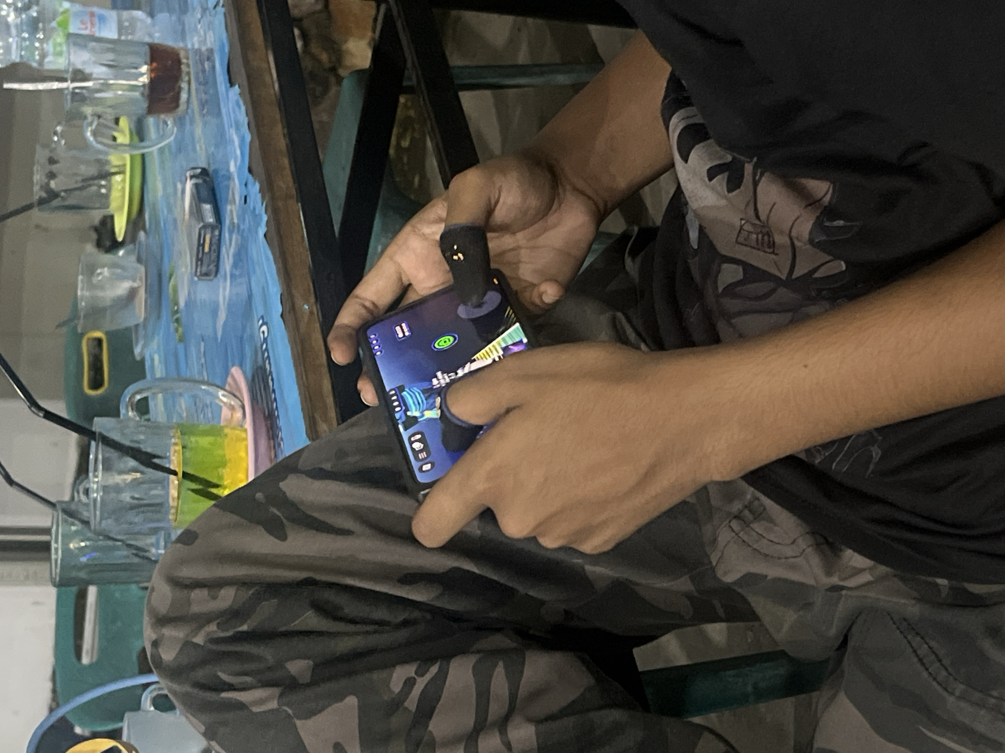 A close-up, vertical shot shows a person playing a mobile game. The person is wearing camouflage pants and a black shirt, partially obscured. Their hands are holding a smartphone, and one thumb is wearing a black gaming sleeve. The phone screen displays a colorful game interface with a circular icon in the center. In the background, out of focus, are several glasses on a blue and white patterned table. One glass contains a yellow liquid with a black straw, another has a dark red liquid, and two are empty. A pack of cigarettes is also visible on the table. The overall setting appears to be an outdoor cafe or restaurant. The image does not provide any specific visual cues to determine the location in Banda Aceh, Indonesia.
