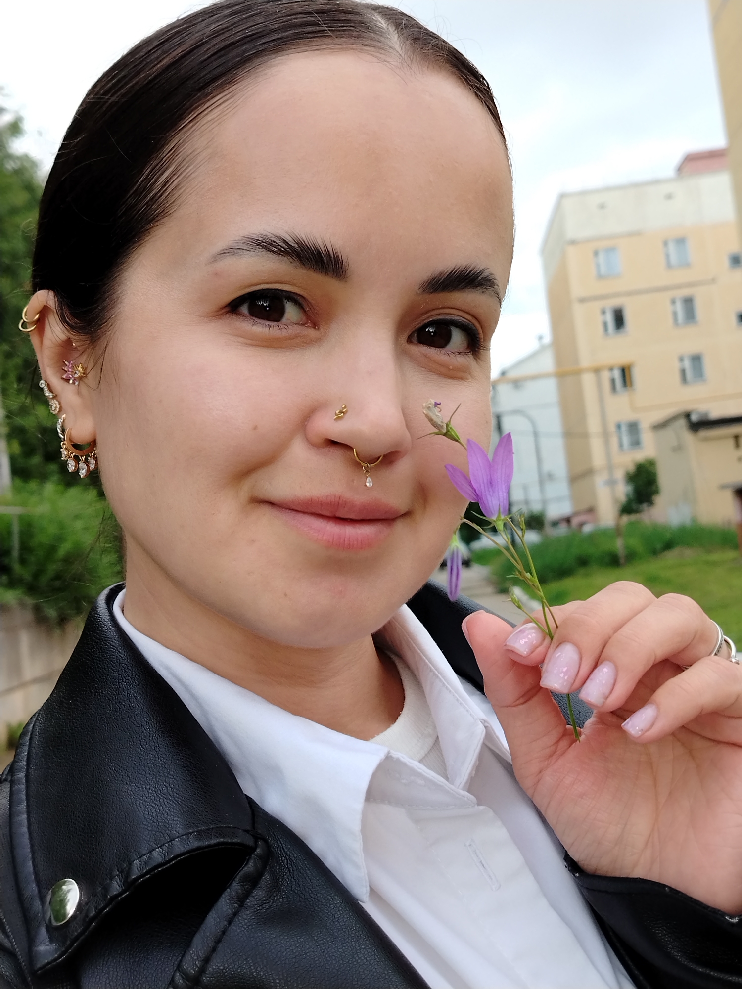 A young adult female is captured in a close-up outdoor shot in Noviy, Russia, looking directly at the camera with a gentle smile. She has dark hair slicked back, dark brown eyes, and smooth skin. She wears a white collared shirt beneath a black faux leather jacket with a visible silver snap button on the lapel.
Her left ear (viewer's left) displays multiple gold-toned piercings: two simple hoops in the upper helix, a pink floral stud in another helix piercing, a conch-like piercing with a cluster of clear stones, and a lobe hoop with multiple dangling clear stones. Her nose features two gold-toned piercings: a small coiled-snake stud in the left nostril and a septum ring with a dangling clear teardrop-shaped stone.
She holds a single purple bell-shaped flower with a slender green stem and one closed bud in her left hand. Her fingernails are manicured short with a light pink, possibly iridescent or glitter-speckled polish. A silver-toned ring is visible on the ring finger of her left hand.
The background, largely out of focus, reveals an overcast sky and a multi-story, light beige or pale yellow building with multiple rectangular windows on the right side. Green foliage is visible behind the subject's left shoulder, suggesting an urban or suburban environment with some natural elements.