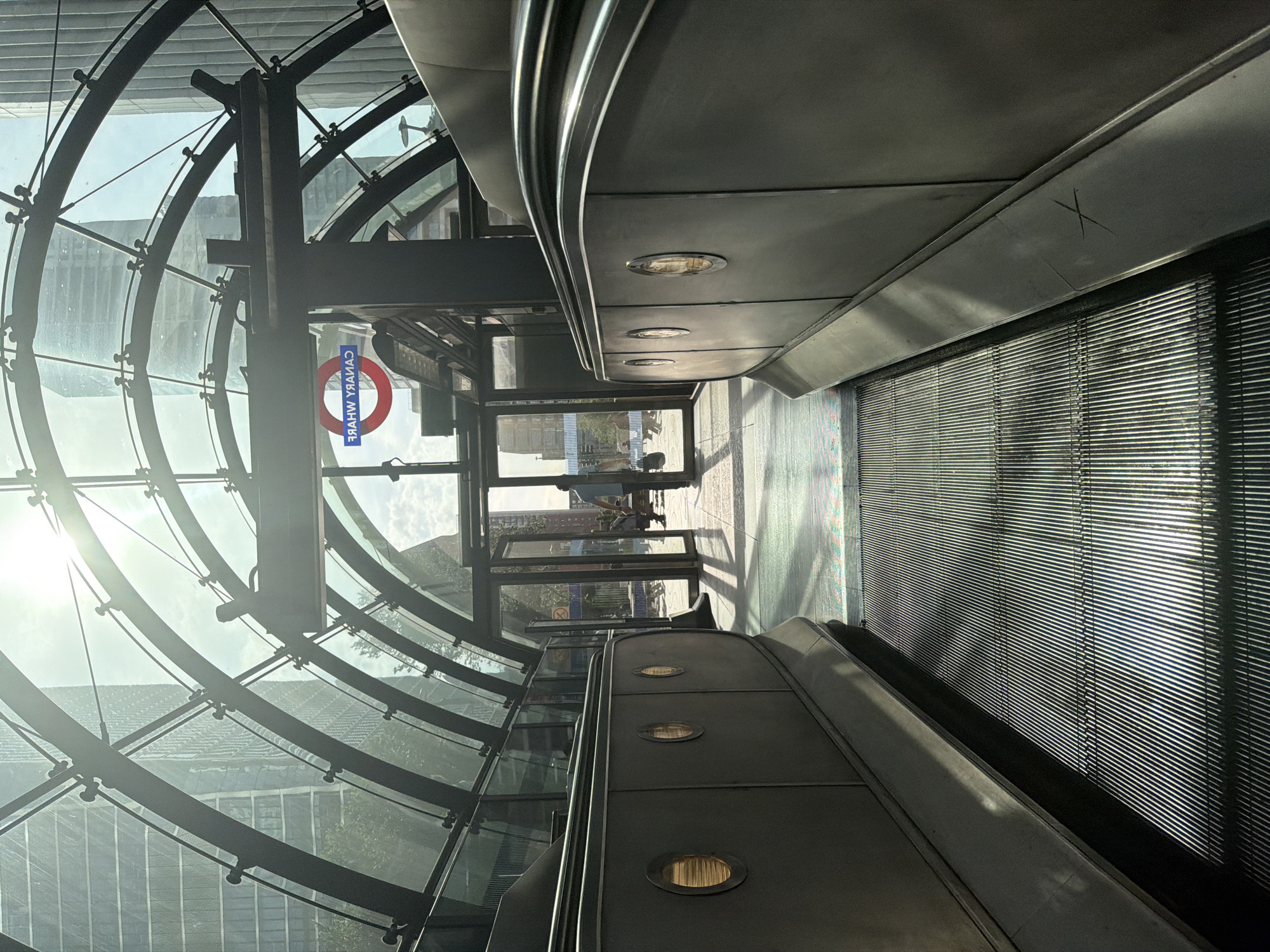 The image depicts an interior view of the Canary Wharf underground station in London, United Kingdom. An escalator ascends towards the platform level, enclosed within a glass and metal structure. The station's signage is visible, indicating the "Central Line." Outside the station entrance, pedestrians are visible in the daylight. The sun is shining brightly through the glass panels. The image is oriented vertically.