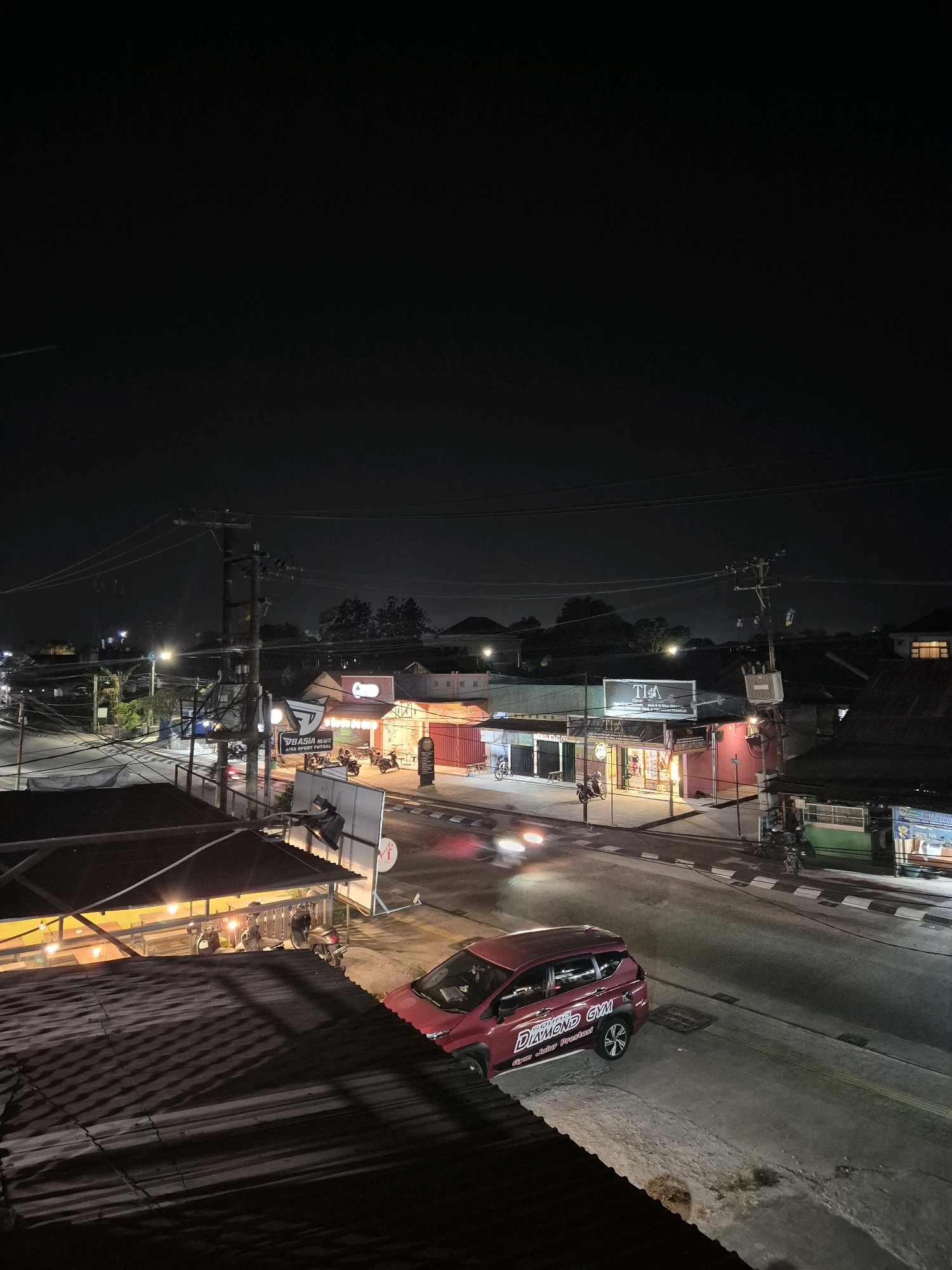 The image depicts a nighttime scene in Bontang, Indonesia, focusing on a street lined with businesses and residential structures. A red SUV, bearing the text "Diamond Gym" and "Gyar Jalar Prestasi", is parked on the side of the road. Several motorcycles are visible in front of illuminated shops. The street is paved and appears to be a main thoroughfare, with streetlights and utility poles casting shadows. The architecture suggests a mix of commercial and residential buildings, with corrugated metal roofs and awnings prominent in the foreground. The overall atmosphere is that of a typical urban street at night, with activity centered around the illuminated storefronts.
