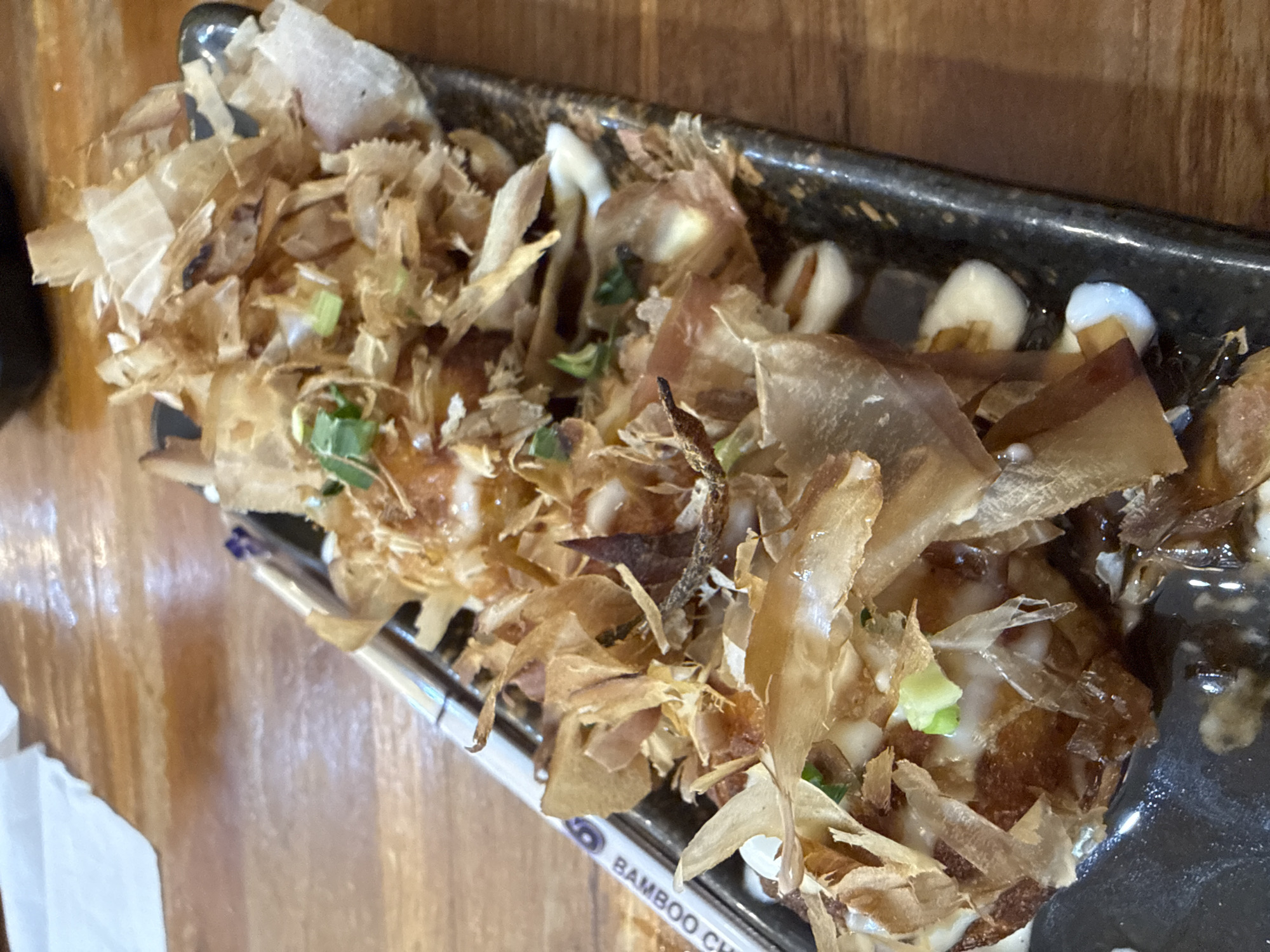 A rectangular black plate of takoyaki is presented at an angle on a wooden surface. The takoyaki is topped with generous amounts of katsuobushi flakes and green onions and what appears to be mayonnaise. A portion of the plate is wet with sauce. A pair of chopsticks labeled "BAMBOO CHI" sits beneath the plate. A partial view of a white napkin is visible on the left side of the frame. The location is unknown to be Lhokseumawe, Indonesia.